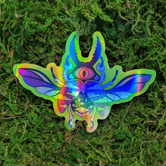 A photograph of a vinyl sticker being held in front of a clematis plant. The vinyl sticker is of a holographic beetle with one eye.