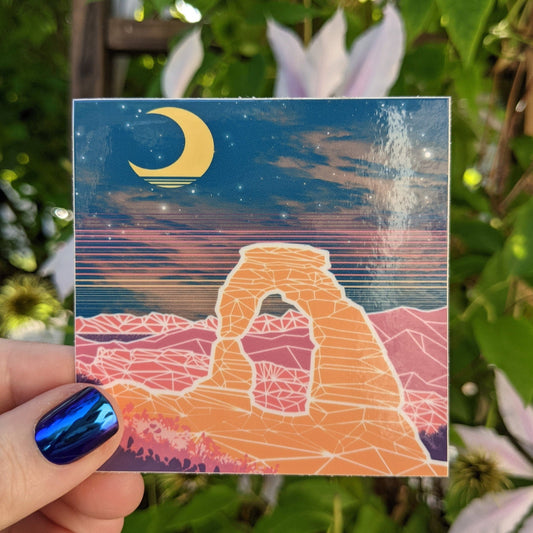 A photograph of a vinyl sticker being held in front of a clematis plant. The vinyl sticker is of delicate arch at Arches national park at night with a vaporwave overlay.