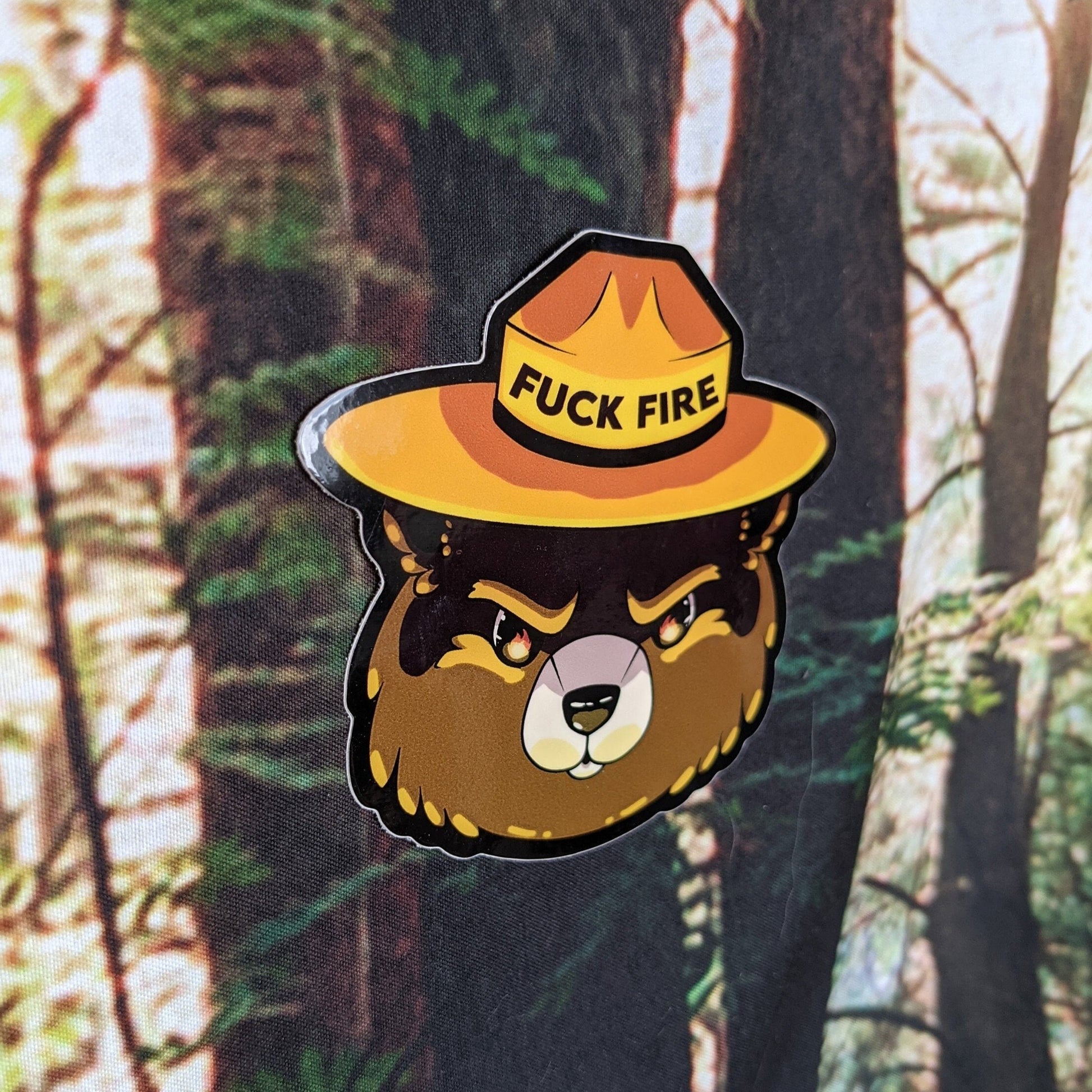 A photograph of a vinyl sticker being held in front of a clematis plant. The sticker is of Smoky the Bear with the reflection of flames in his eyes. His ranger hat says, “Fuck fire.”