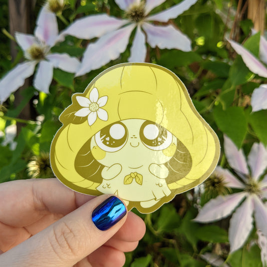 A photograph of a vinyl sticker being held in front of a clematis plant. The sticker is of a yellow mushroom sprite called a Mushkin.
