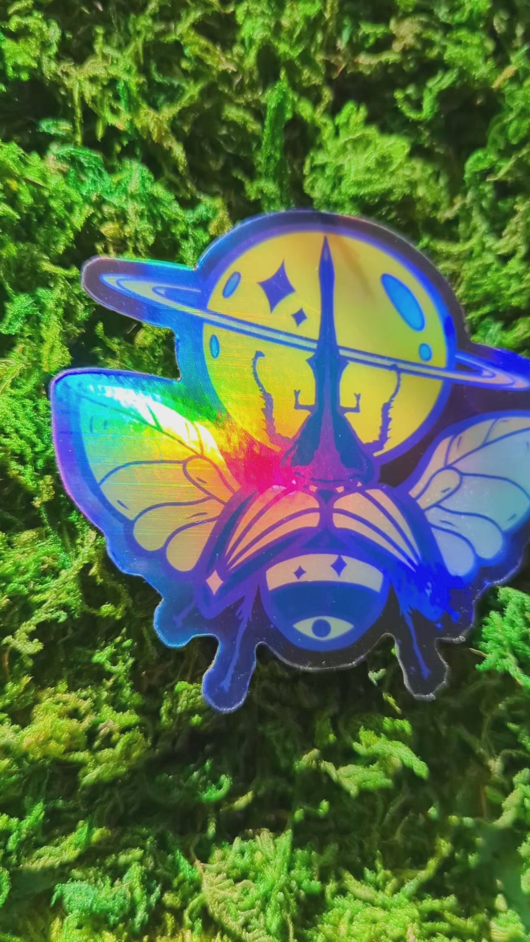 Video of a mossy background with a holographic vinyl sticker of a saturn themed beetle. The video shows how shiny the holographic effect is.