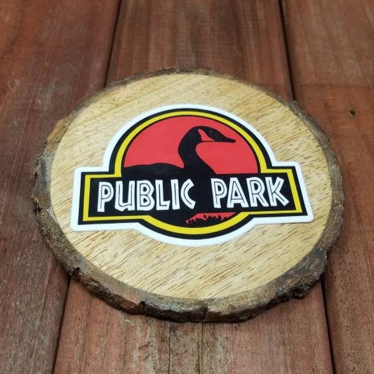 Photograph of a red wood surface with a wood round. On top of the wood round is a vinyl sticker of a goose in front of “Public park” in a parody style to Jurassic Park.