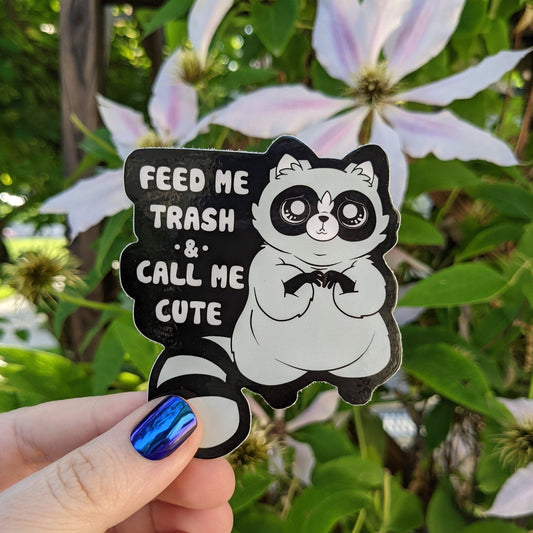 A photograph of a vinyl sticker being held in front of a clematis plant. The vinyl sticker is a raccoon making sad eyes and begging. To the left of the raccoon it says, “Feed me trash & call me cute.”