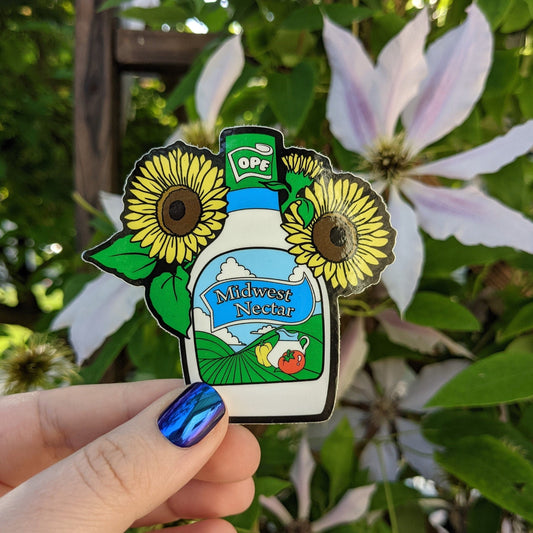 A photograph of a vinyl sticker being held in front of a clematis plant. The vinyl sticker is a bottle of ranch surrounded by sunflowers. The ranch bottle says, “Midwest Nectar.”