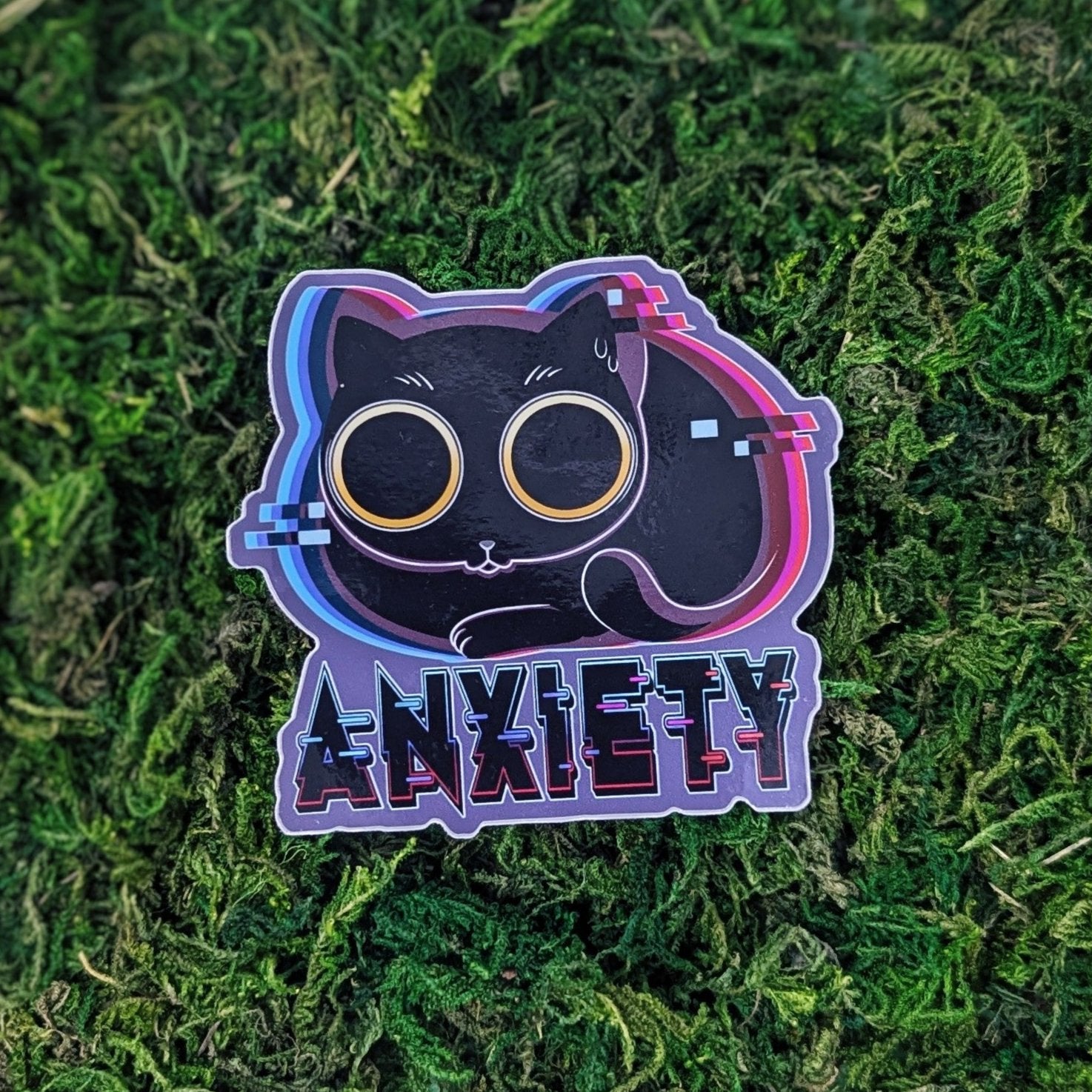 A photograph of a sticker on some moss. The sticker is of a nervous cat with giant eyes glitching out. Under it says, Anxiety.