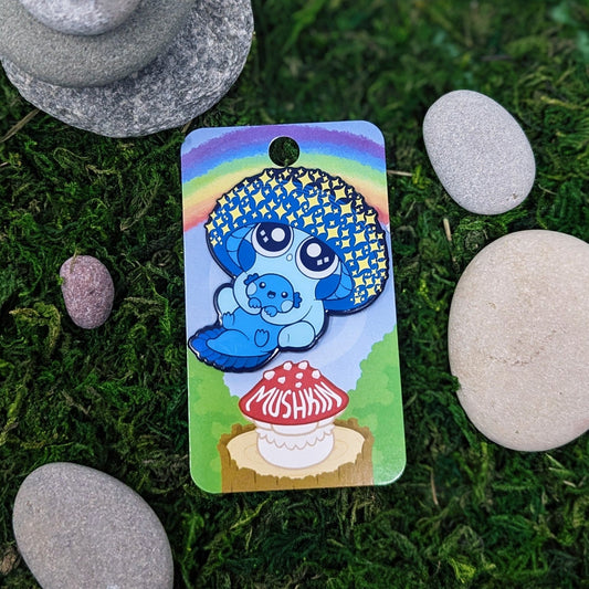 Photograph of a mossy background with an enamel pin of a sparkly blue mushroom sprite called a Mushkin. This Mushkin is holding a little blue axolotl.