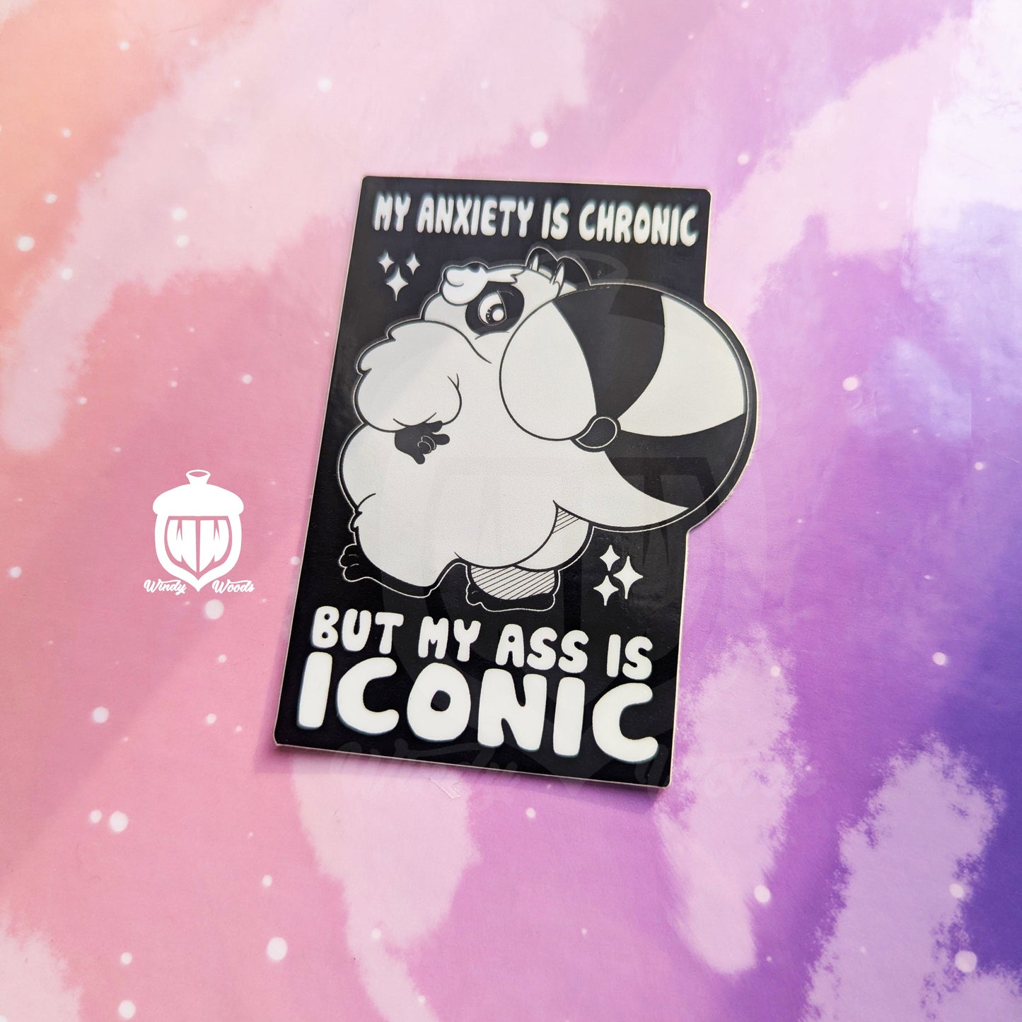 A photograph of a vinyl sticker against a pink and purple background. The sticker features Chaps the Raccoon standing with his little booty out. Around him it says, “My anxiety is chronic but my ass is iconic.”