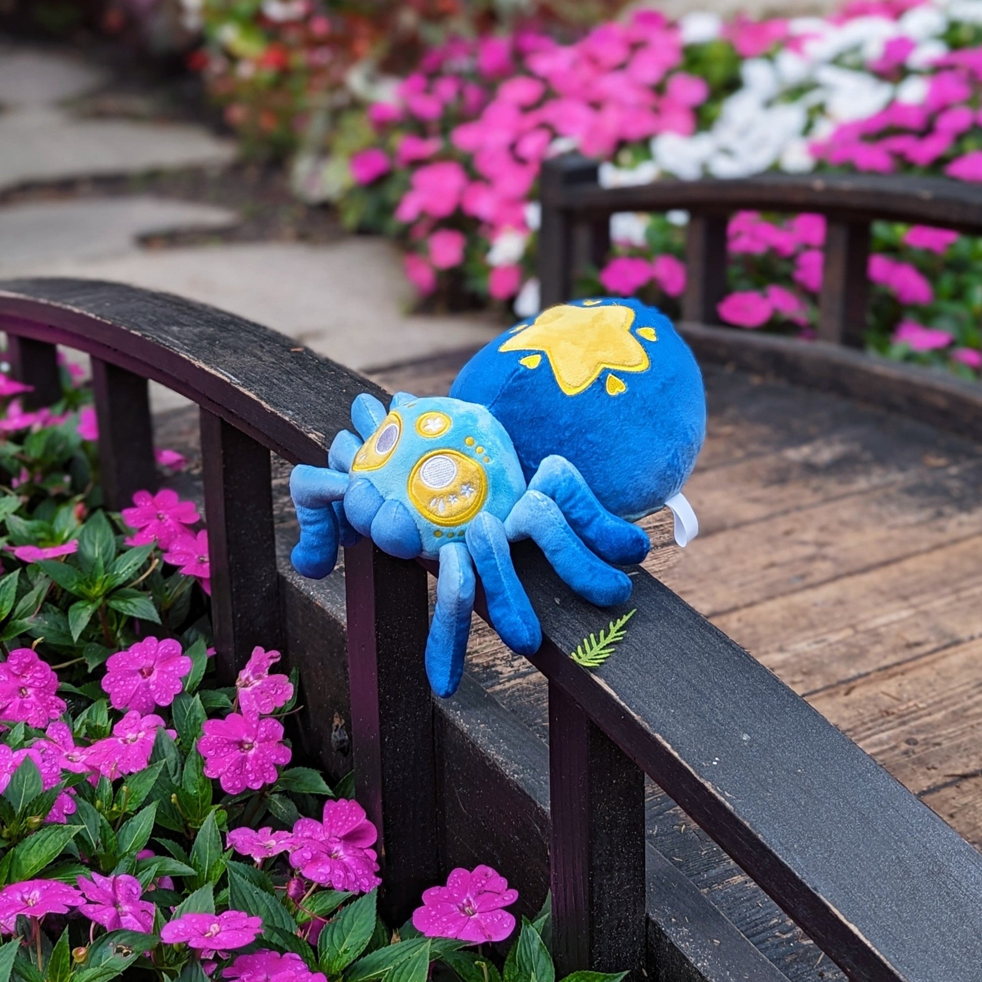 A photograph of bridge over flowers with a plush spider on it. The spider's name is Cosmo. They are blue with yellow eyes and a yellow star on it's butt.