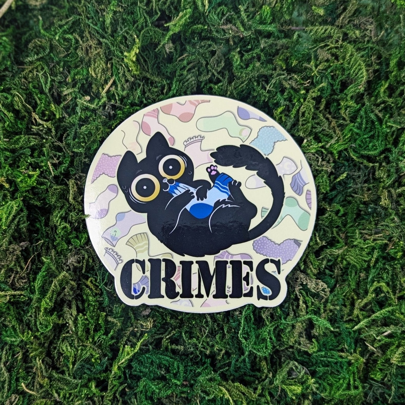 A photograph of a sticker on some moss. The sticker is vinyl and is of a cat chewing on a sock surrounded by socks of different patterns. Under the cat it says, Crimes.