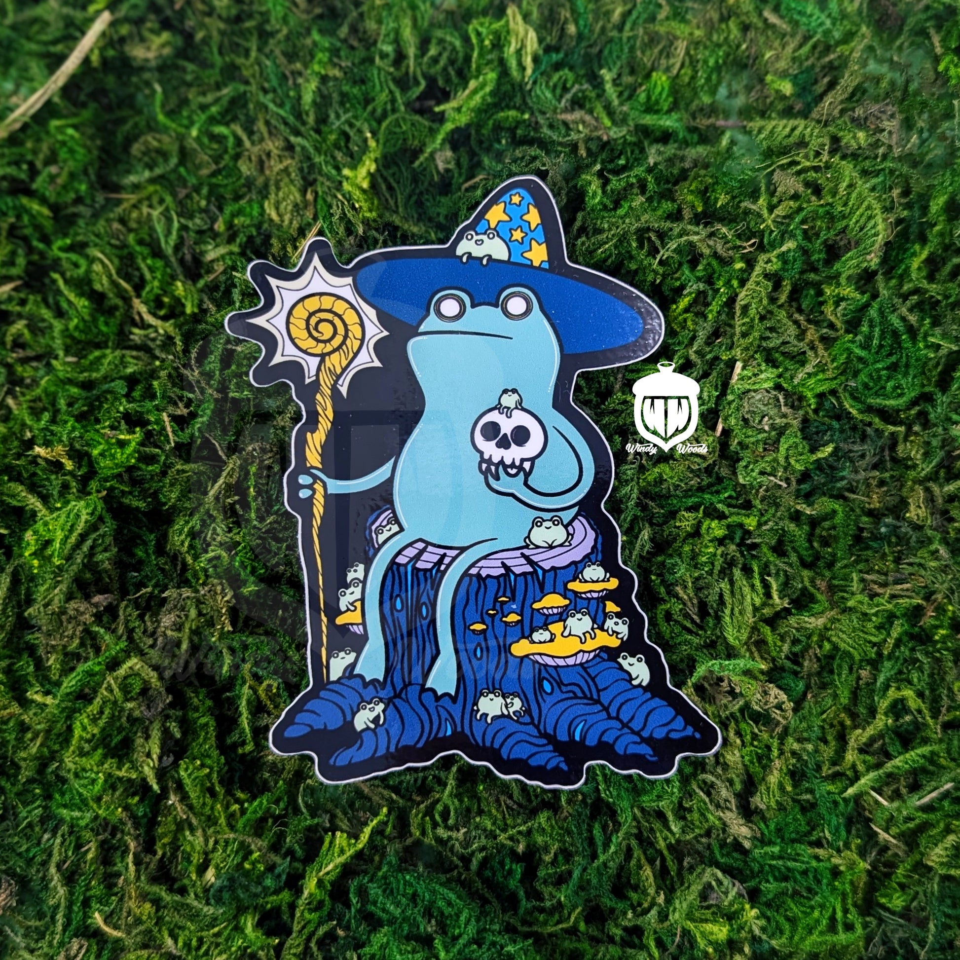 A Photograph of a sticker on some moss. The sticker features artwork of a bipedal green frog sitting on a stump. This is the Loveland Frogman Cryptid. The Frogman is wearing a blue wizard hat with yellow stars on it. They're holding a yellow branch staff that's glowing with power. The Frogman is holding a skull in their other hand. They are covered and surrounded by small frogs that they definitely summoned to their side.