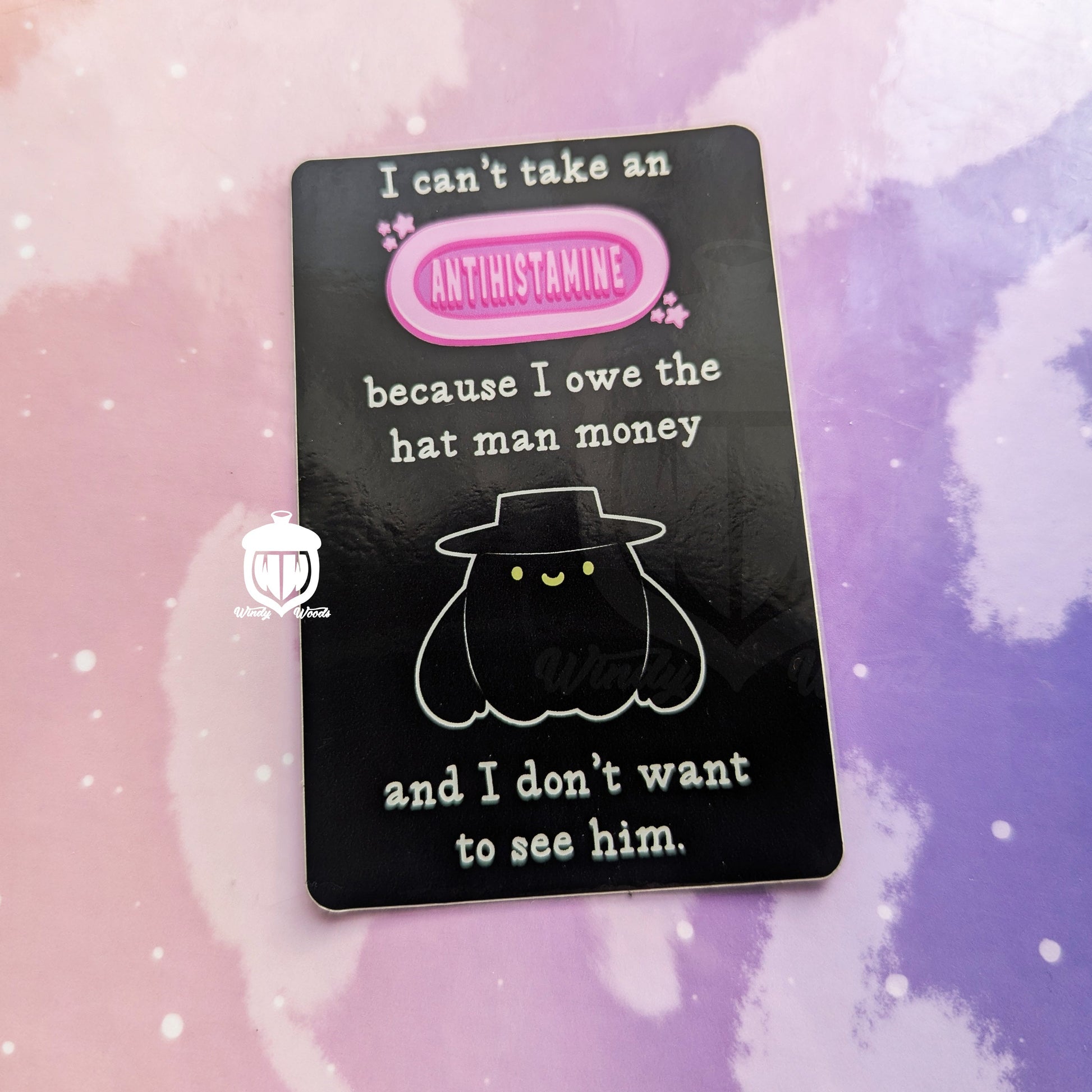 A photograph of a vinyl sticker against a pink and purple background. The sticker features the cryptid, the Hat Man. Around him it says, “I can’t take an antihistamine because I owe the Hatman money and I don’t want to see him.”