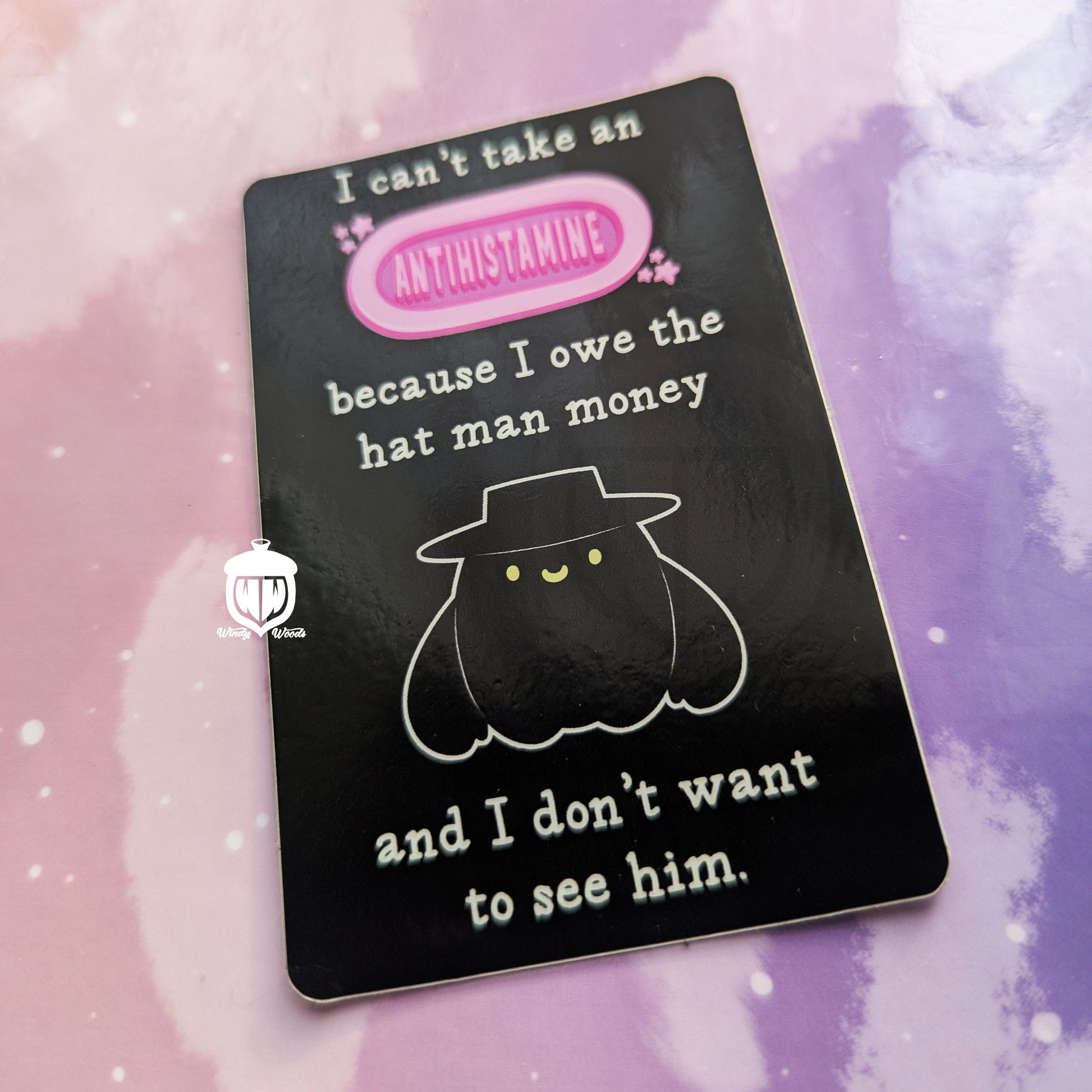 A photograph of a vinyl sticker against a pink and purple background. The sticker features the cryptid, the Hat Man. Around him it says, “I can’t take an antihistamine because I owe the Hatman money and I don’t want to see him.”