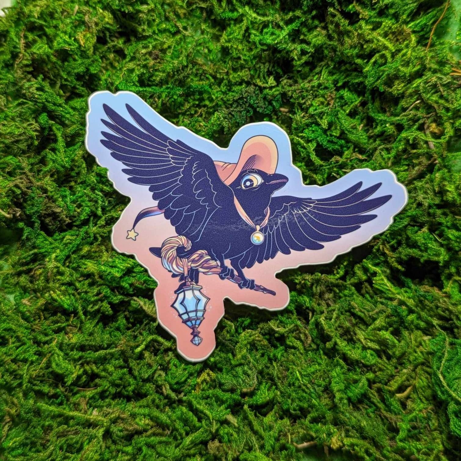 Photograph of a mossy background with a vinyl sticker of Ikarus the Raven Witch in their orange witch hat. Ikarus is carrying in a wood staff with a blue lantern hanging from the hook.