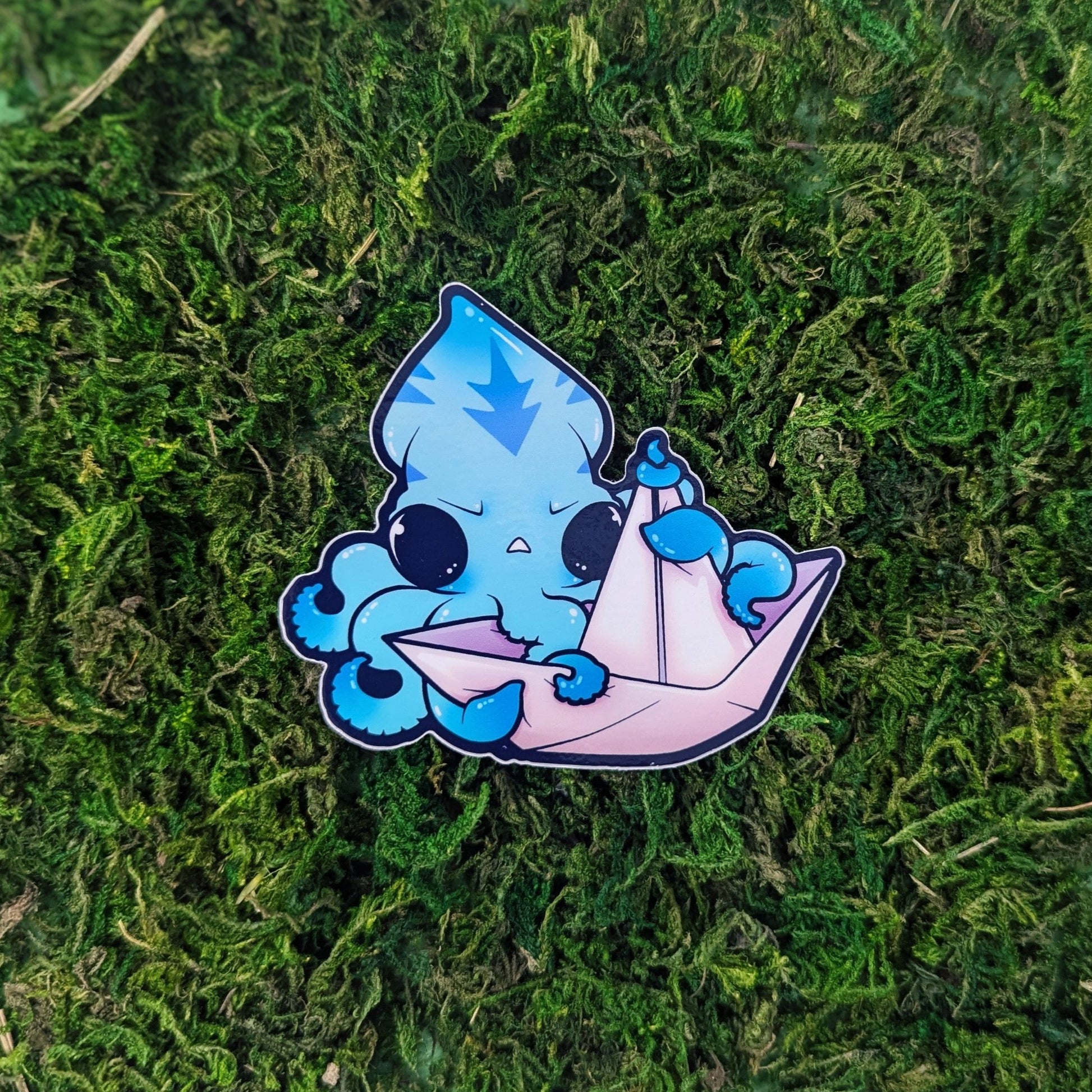 A photograph of an enamel pin light blue squid grabbing onto a paper boat on a mossy background.