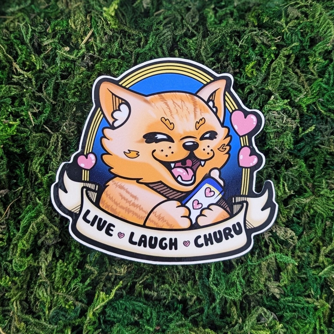 A photograph of a sticker on some moss. The sticker is of an orange cat holding a liquid cat treat with hearts around it. Below it says, "Live Laugh Churu"