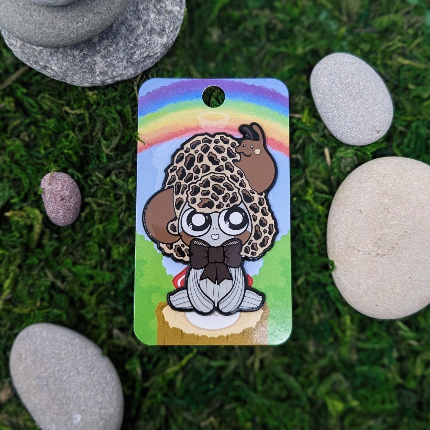 Photograph of a mossy background with an enamel pin of a Morel Mushroom Sprite called a Mushkin. The Mushkin is sitting and looking up at a slug coming out of it’s cap.