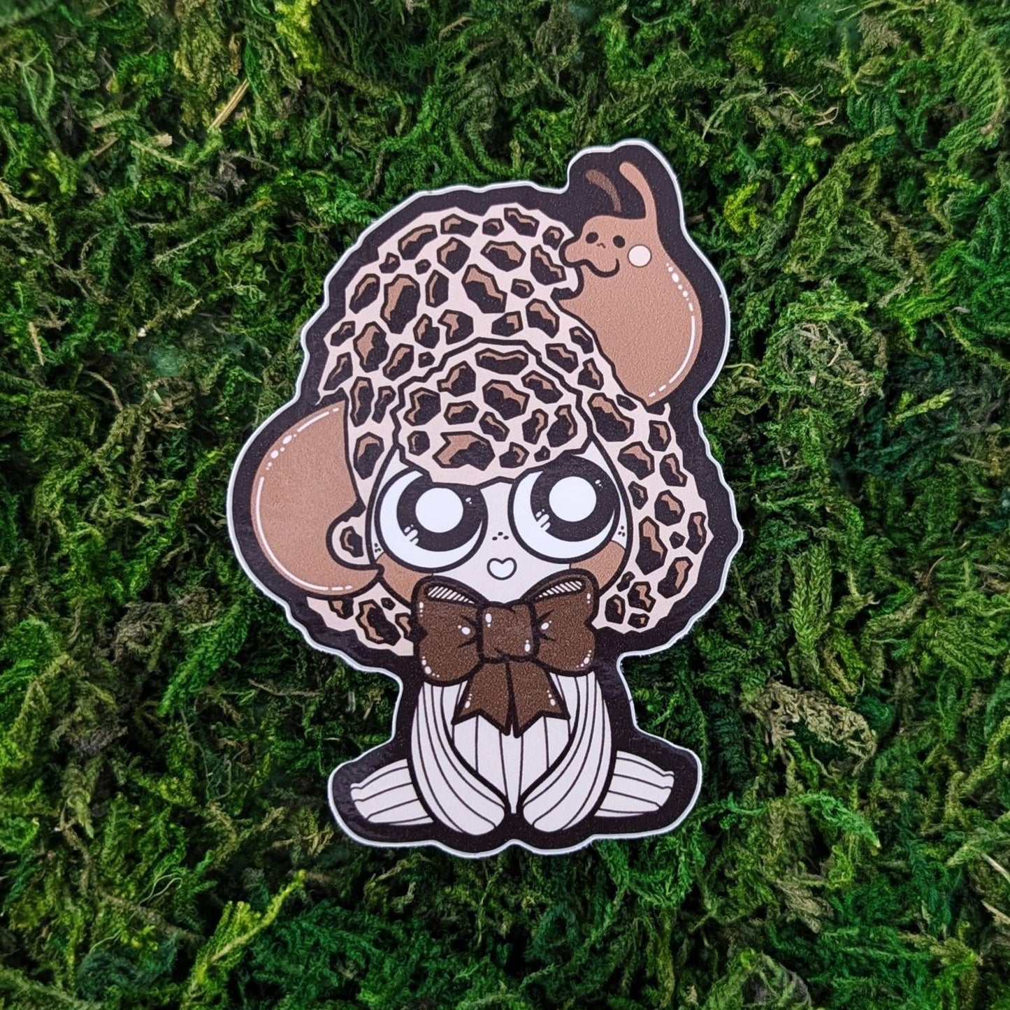 Photograph of a mossy background with a vinyl sticker of a Morel themed Mushkin with a slug coming out of it’s head.