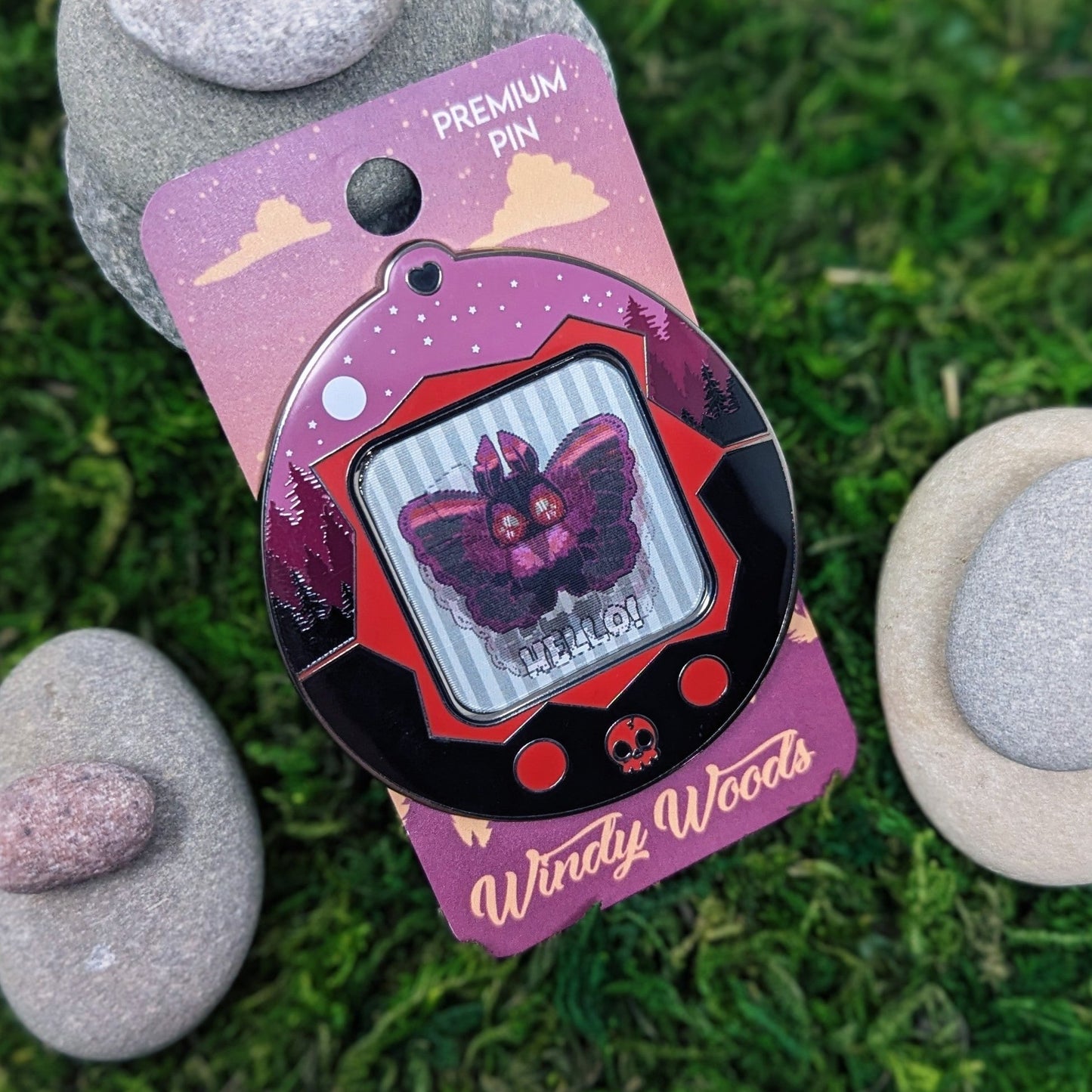 Photograph of a mossy background with an enamel pin of a a pink and black Mothman. The Mothman is in a tamagotchi type device that is a red and black forest. The screen of the tamagotchi is lenticular and is animated to show Mothman saying hello.