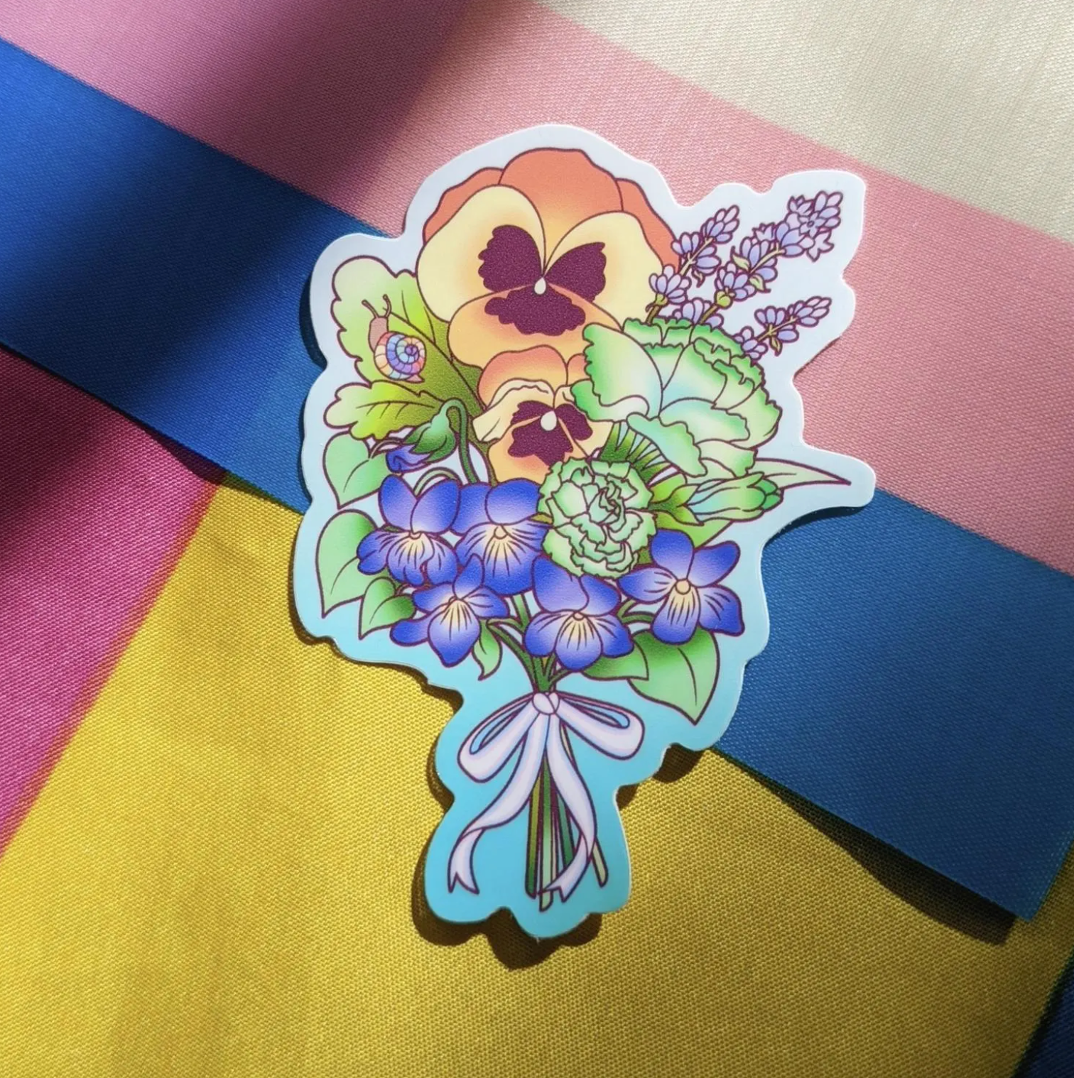 A photograph of a background with the transgender and pansexual pride flags. On top of the flags is a vinyl sticker of a flower bouquet featuring queer themes.