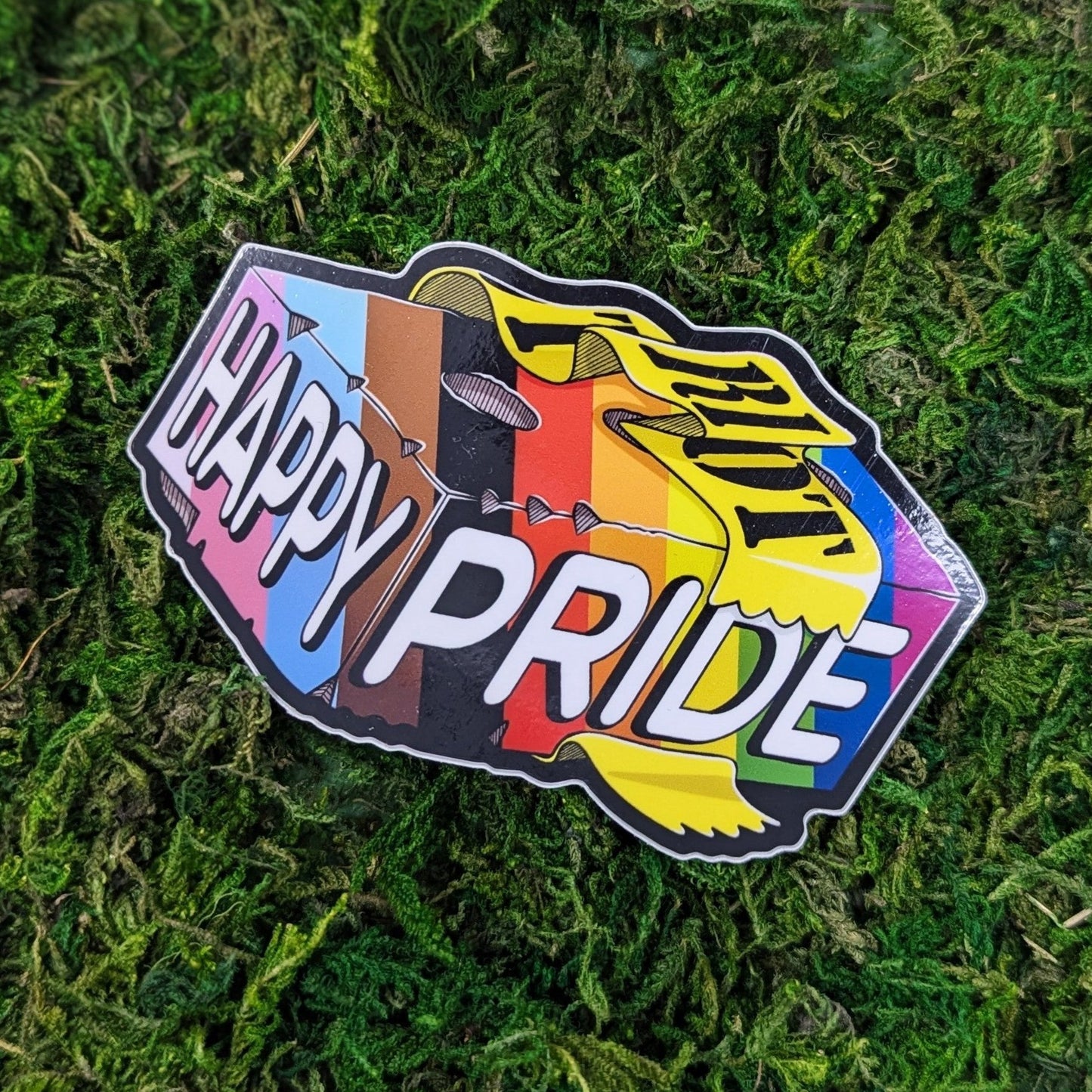 Photograph of a mossy background with a vinyl sticker of a brick in progress flat pride colors that says, “Happy Pride” with a piece of caution tape around it that says, “Riot.”