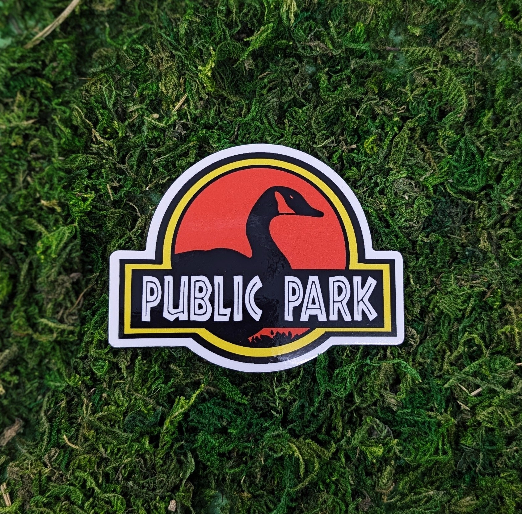 Photograph of a mossy background with a vinyl sticker of a goose in front of “Public park” in a parody style to Jurassic Park.