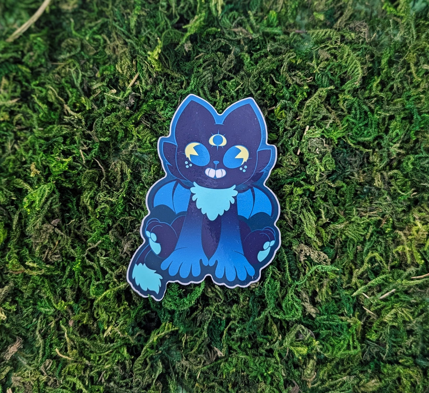 Photograph of a mossy background with a vinyl sticker of Rigel the shadow cat cryptid. Rigel is dark blue with yellow and teal eyes and a third eye on his forehead.