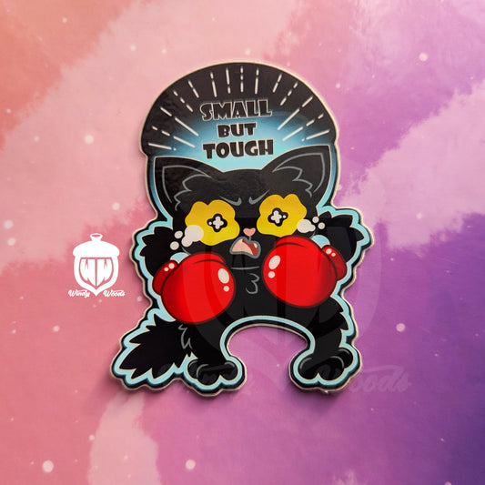 A photograph of a vinyl sticker against a pink and purple background. The sticker features a small black cat with red boxing gloves. The cat is crying with a furrowed brow. Above the cat it says, “Small but tough.”