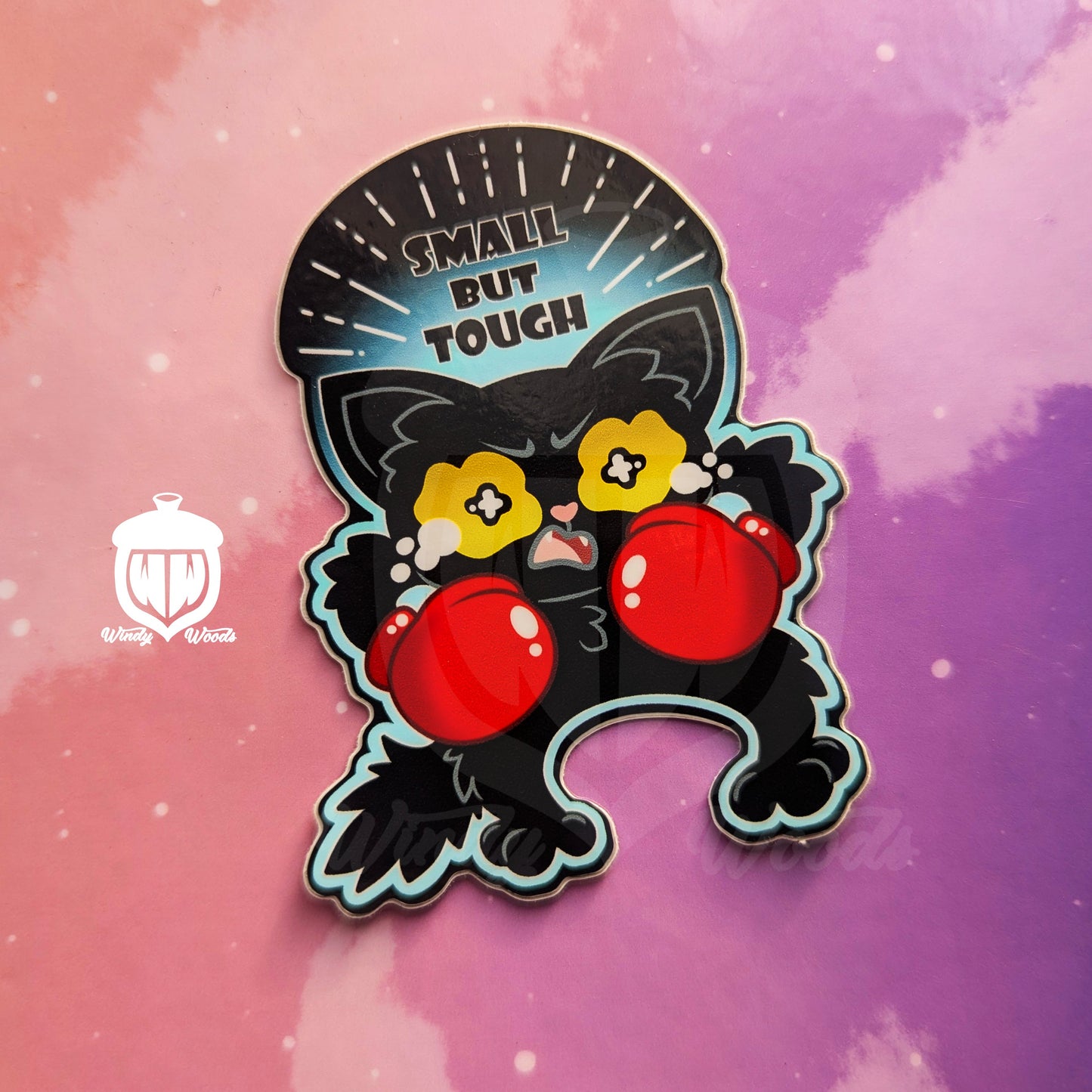 A photograph of a vinyl sticker against a pink and purple background. The sticker features a small black cat with red boxing gloves. The cat is crying with a furrowed brow. Above the cat it says, “Small but tough.”