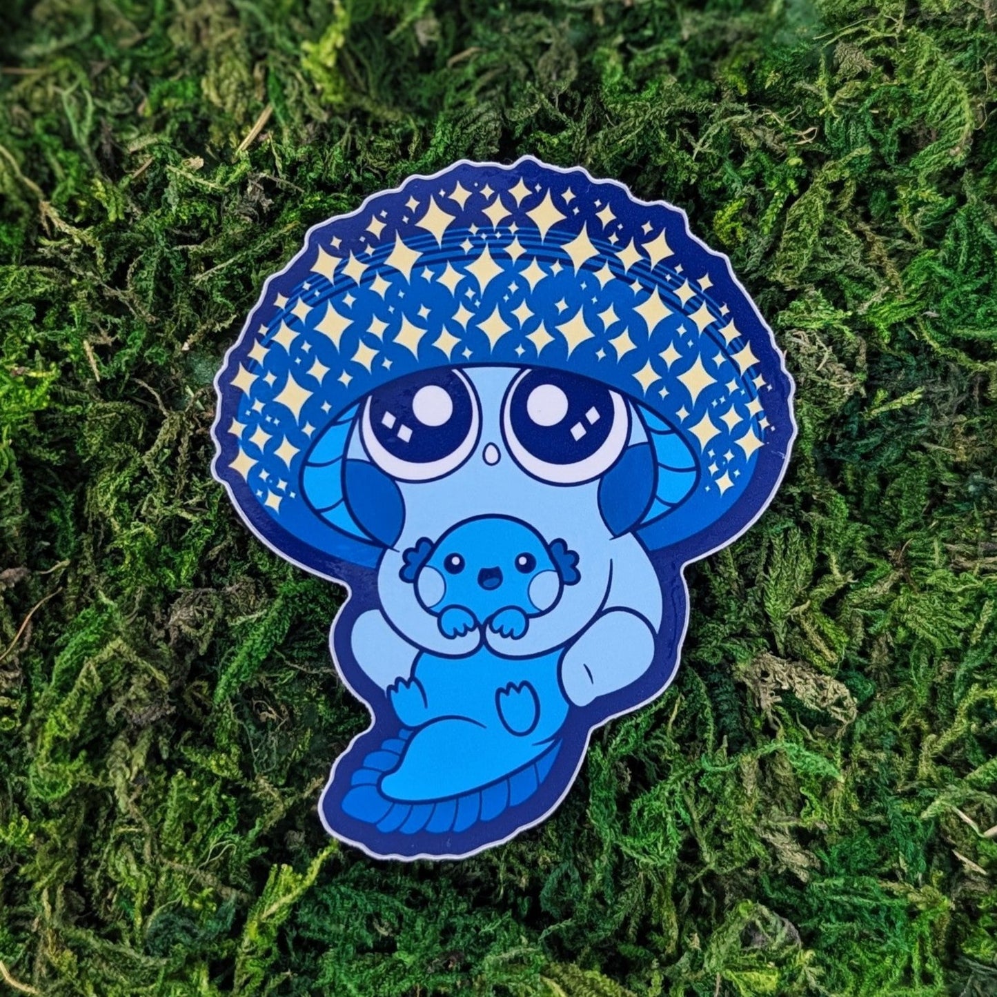 Photograph of a mossy background with a vinyl sticker of a Blue Sparkly Mushkin hugging an axolotl and star gazing.