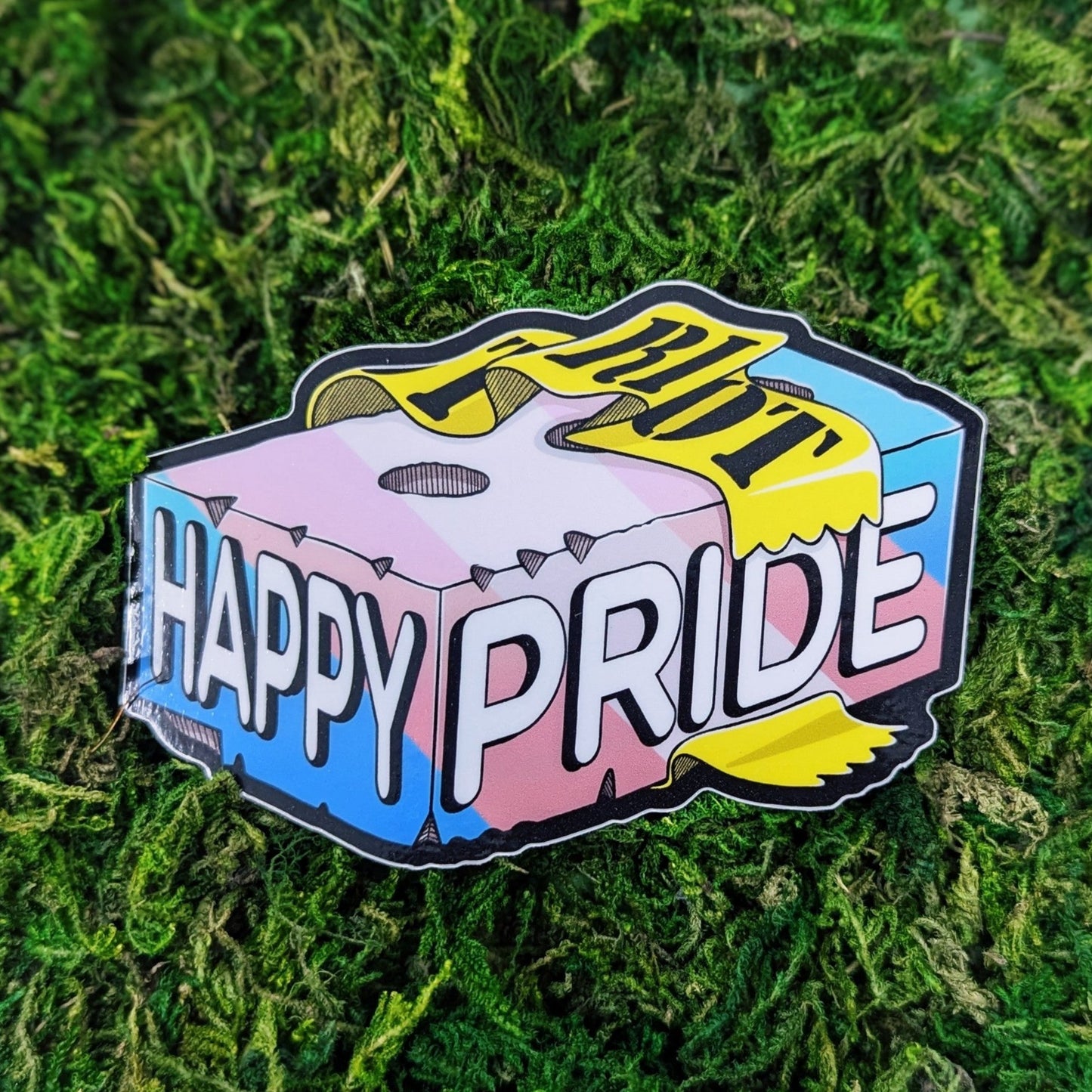 Photograph of a mossy background with a vinyl sticker of a brick in transgender pride colors that says, “Happy Pride” with a piece of caution tape around it that says, “Riot.”