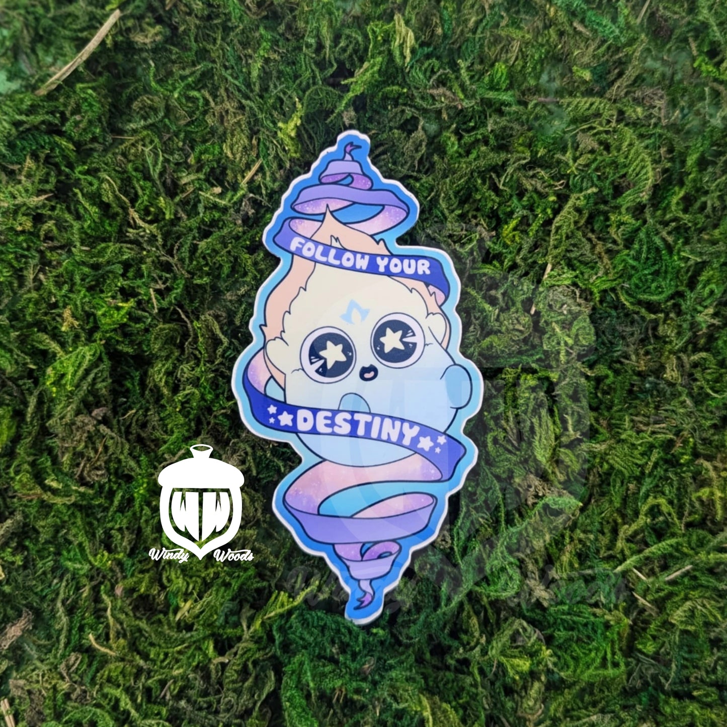 A photograph of a sticker on some moss. The sticker features a Will-o'-the-wisp with stars in its eyes. The flames are pastel yellow, blue, and orange. A scroll around him says, "Follow your Destiny."