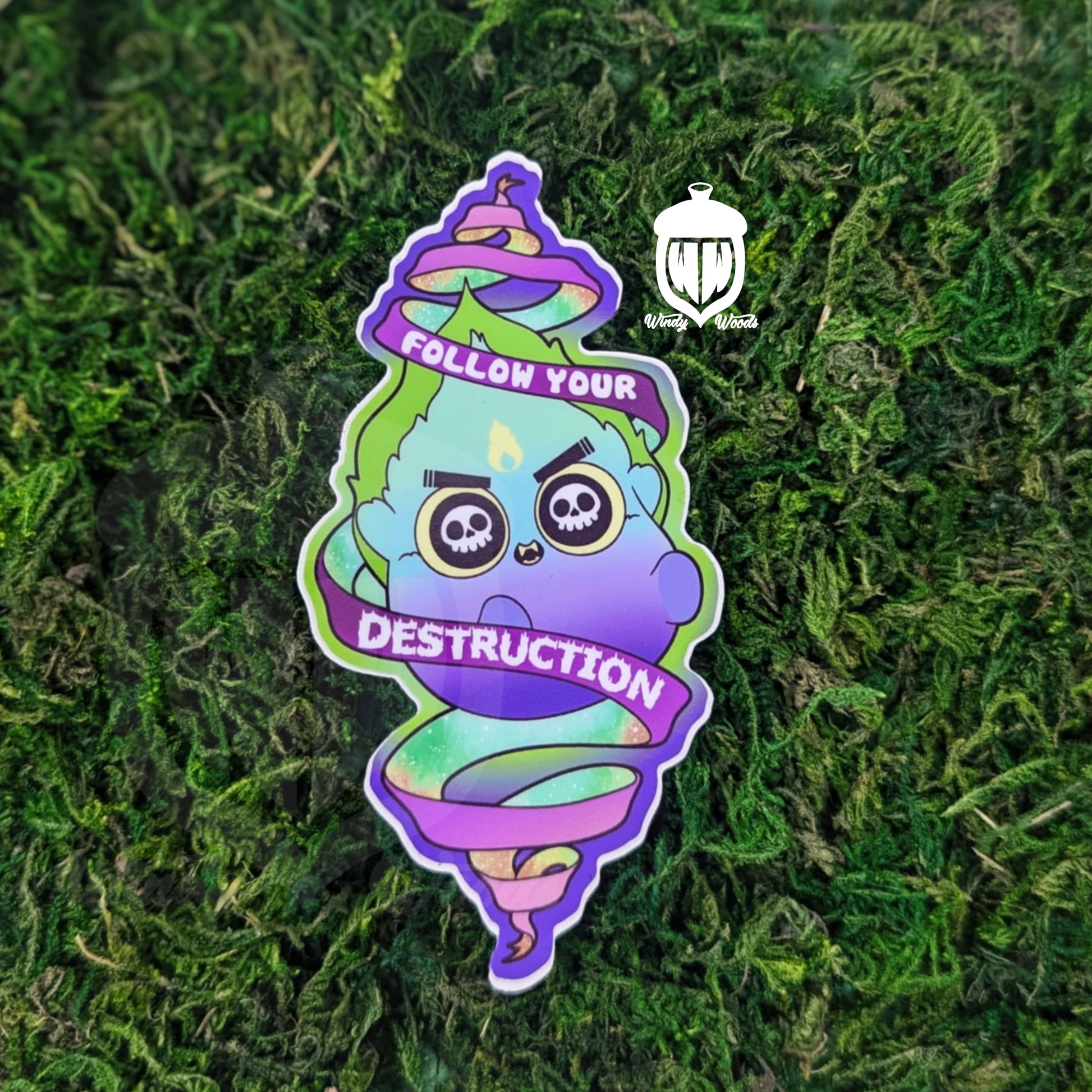 A photograph of a sticker on some moss. The sticker features a Will-o'-the-wisp with skulls in it's eyes. The flames are green blue and purple. A scroll around him says, "Follow your destruction."