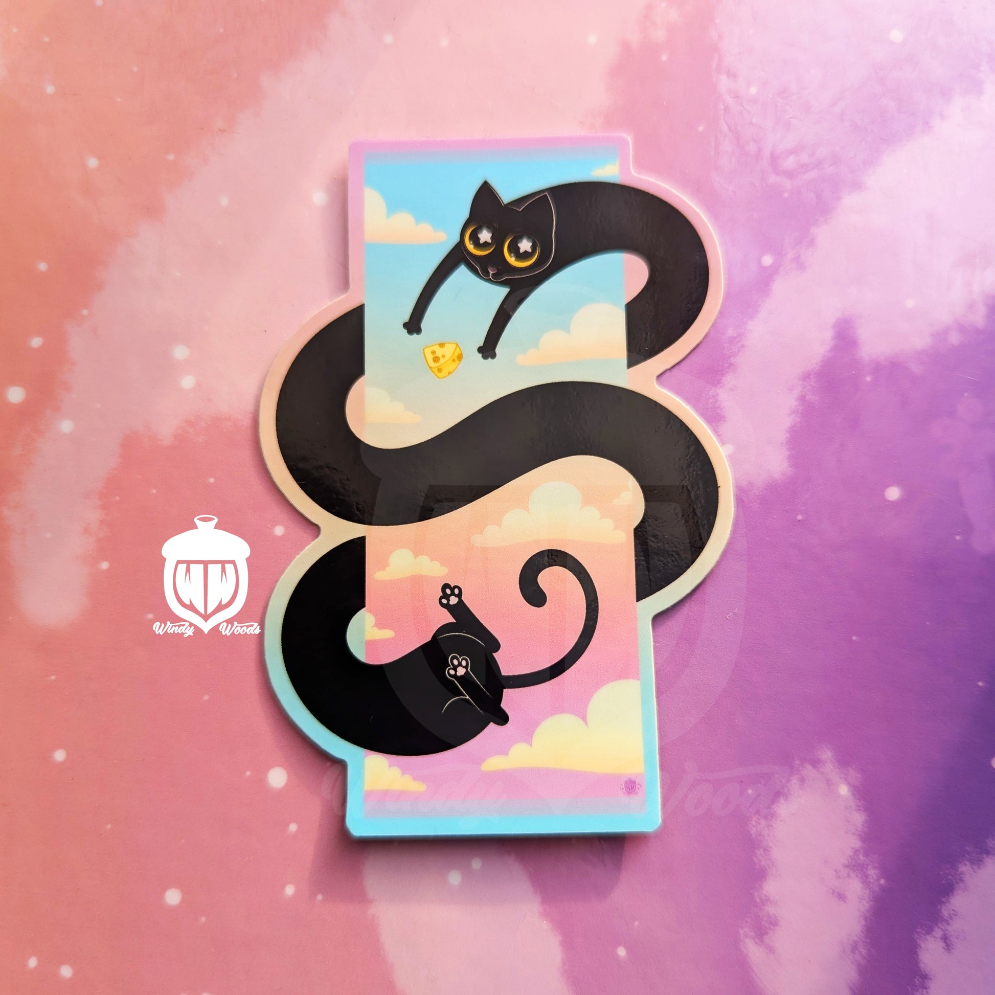 A photograph of a vinyl sticker against a pink and purple background. The sticker features a long black cat swirling in a spiral around a sunset sky. Near their arms is a cat toy that looks like a block of cheese.