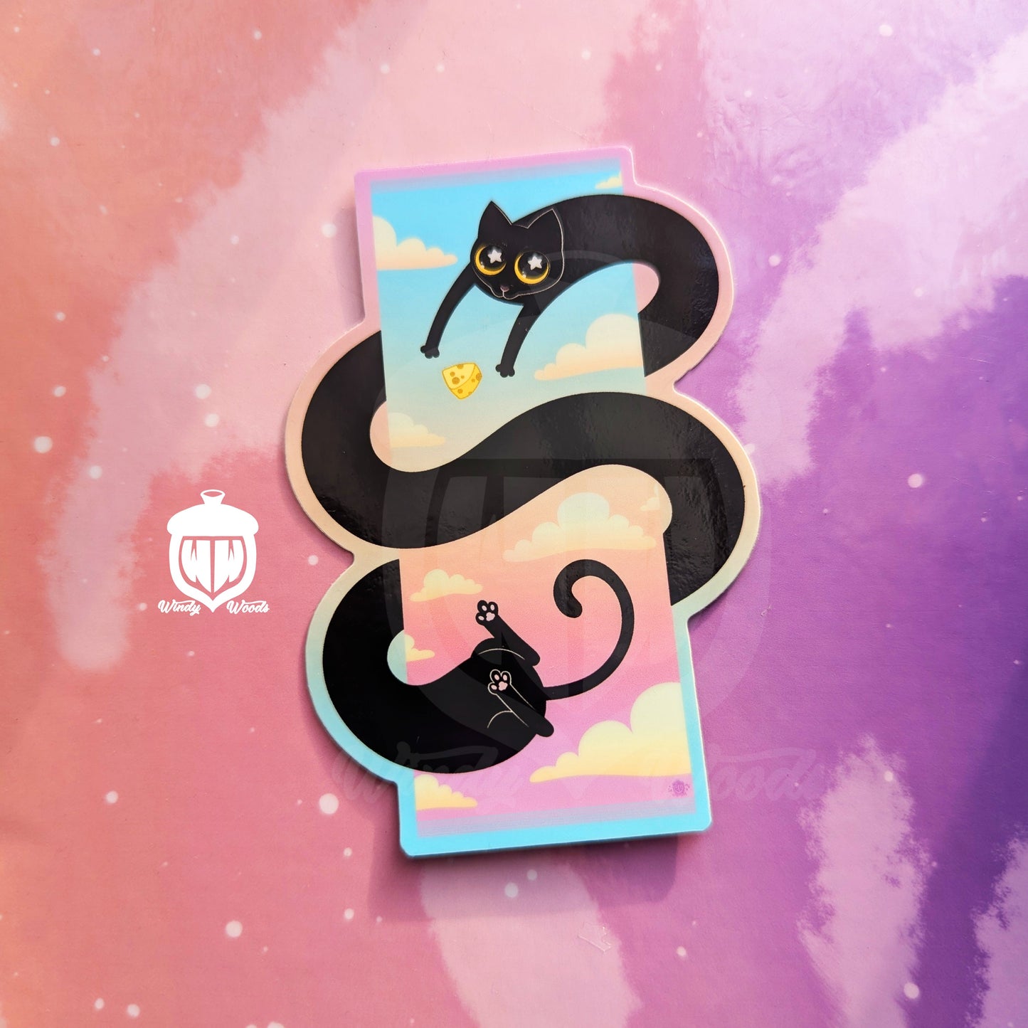 A photograph of a vinyl sticker against a pink and purple background. The sticker features a long black cat swirling in a spiral around a sunset sky. Near their arms is a cat toy that looks like a block of cheese.