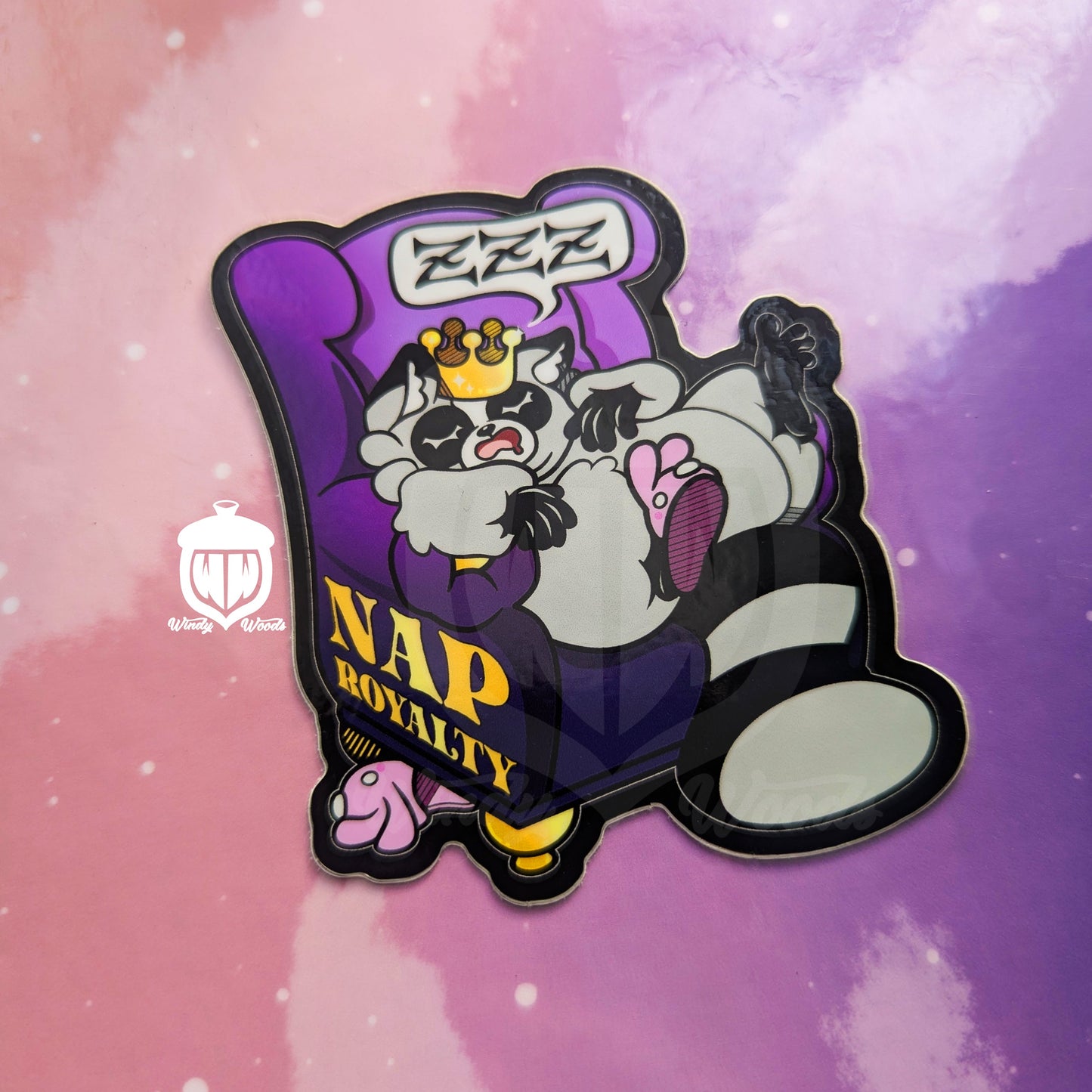 A photograph of a vinyl sticker against a pink and purple background. The sticker features Chaps the Raccoon napping on a purple chair and wearing a crown. On the side of the chair in gold it says, “Nap Royalty.”