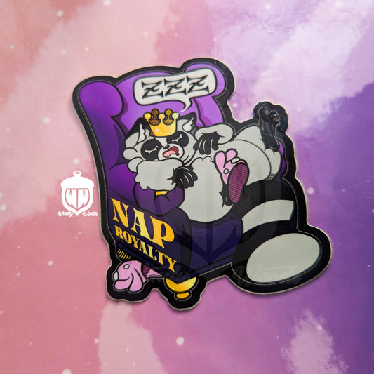A photograph of a vinyl sticker against a pink and purple background. The sticker features Chaps the Raccoon napping on a purple chair and wearing a crown. On the side of the chair in gold it says, “Nap Royalty.”