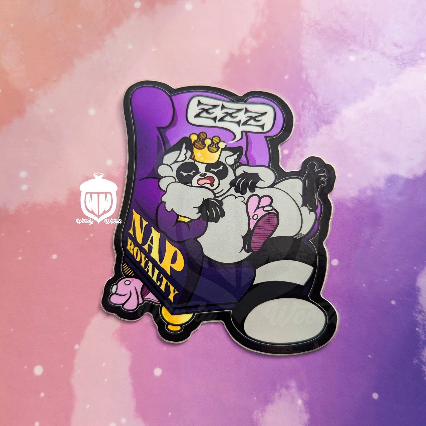 A photograph of a vinyl sticker against a pink and purple background. The sticker features Chaps the Raccoon napping on a purple chair and wearing a crown. On the side of the chair in gold it says, “Nap Royalty.”