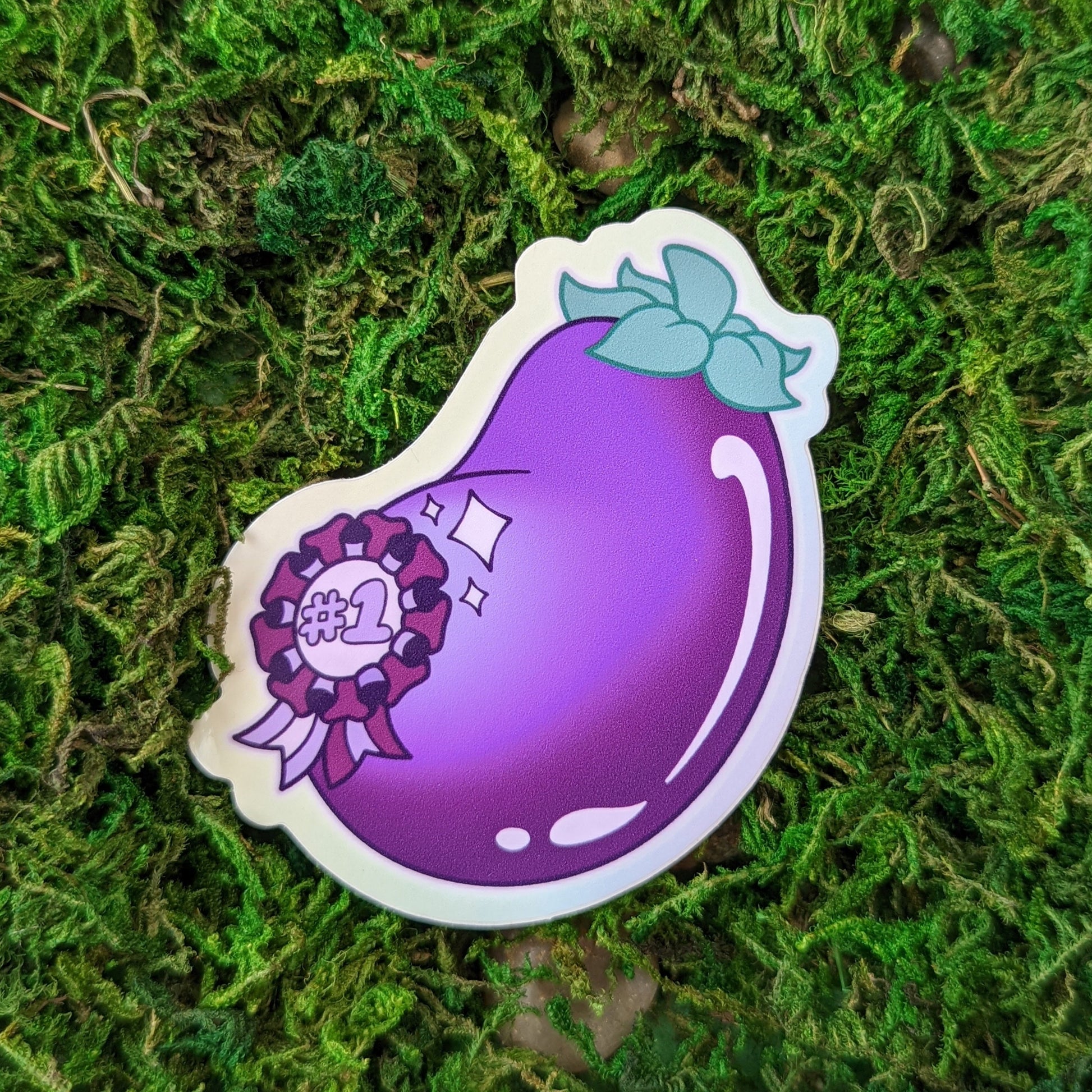 Photograph of a mossy background with a vinyl sticker of an eggplant with a sparkly #1 ribbon on it.