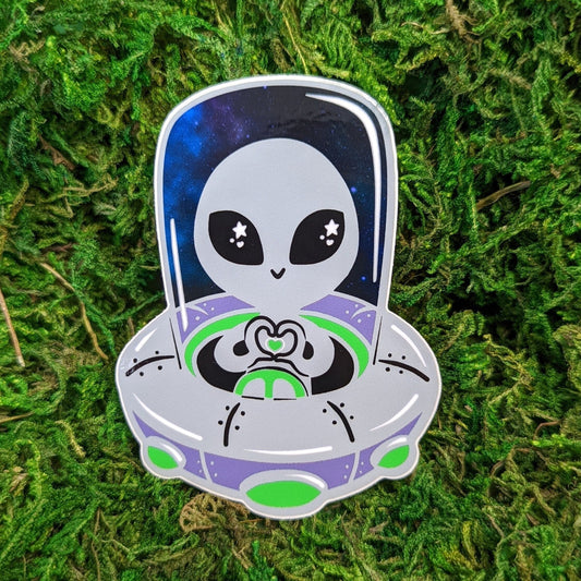 Photograph of a mossy background with a vinyl sticker of a cute alien in a UFO. The gray alien is making a heart shape with its two hands.