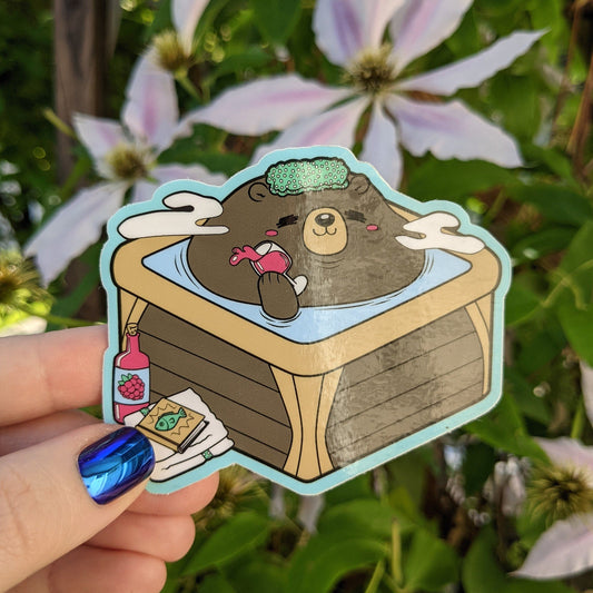 A photograph of a vinyl sticker being held in front of a clematis plant. The sticker is a hot tub with a bear in it enjoying a glass of grape juice.