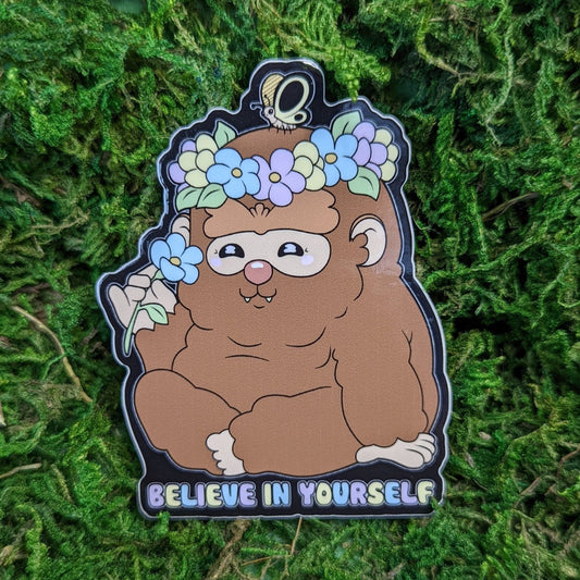 Photograph of a mossy background with a vinyl sticker of Bigfoot sitting with a flower crown and sniffing a flower. Under Bigfoot it says, “Believe in yourself.”