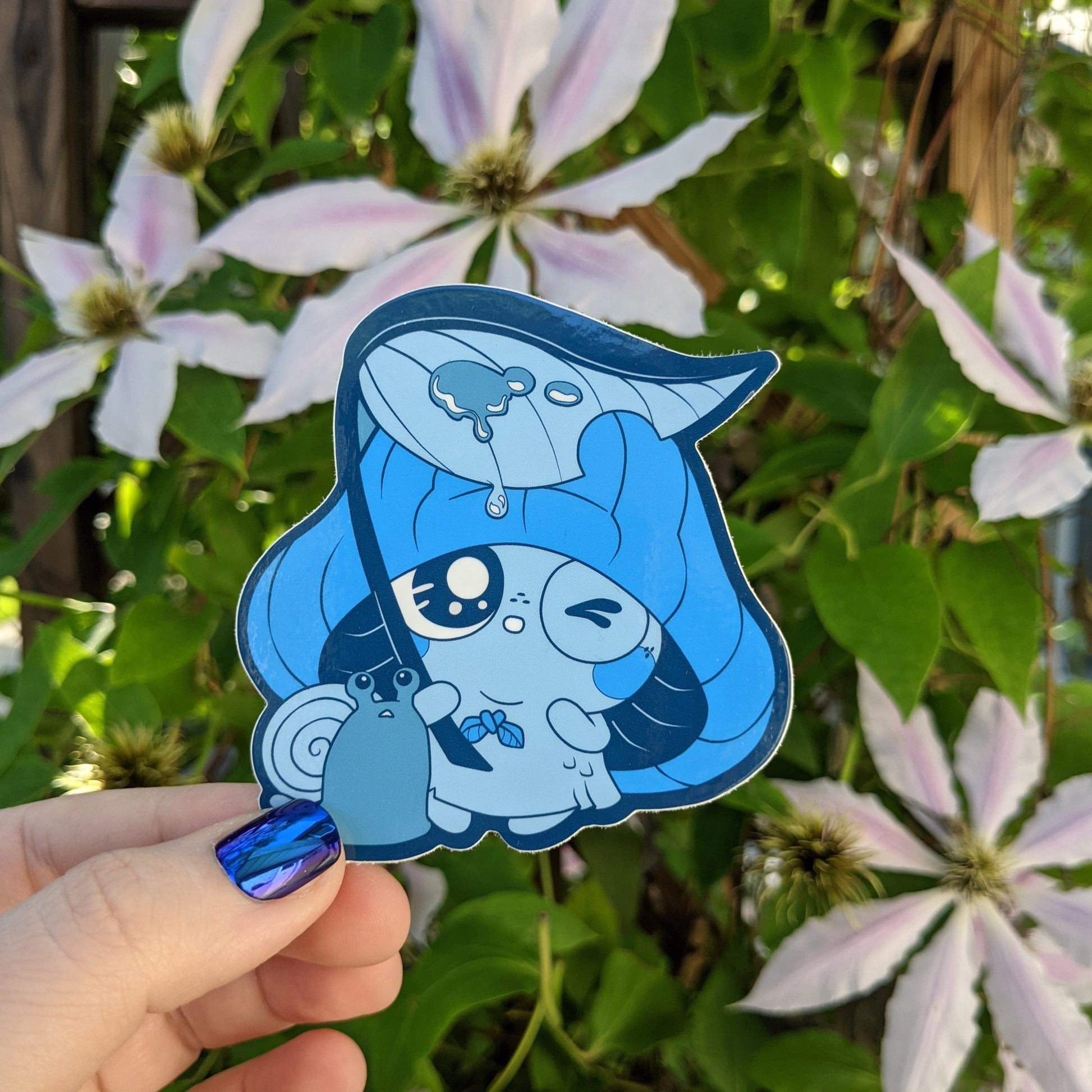 A photograph of a vinyl sticker being held in front of a clematis plant. The sticker is of a blue mushroom sprite called a Mushkin. Mushkin is hiding with a snail under a leaf umbrella in the rain.