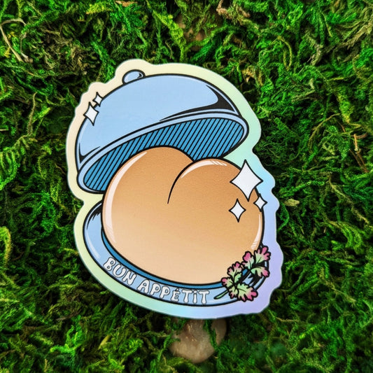 Photograph of a mossy background with a vinyl sticker of a serving tray presenting a booty shaped bread bun. On the bottom plate it says, “Bun Appétit."