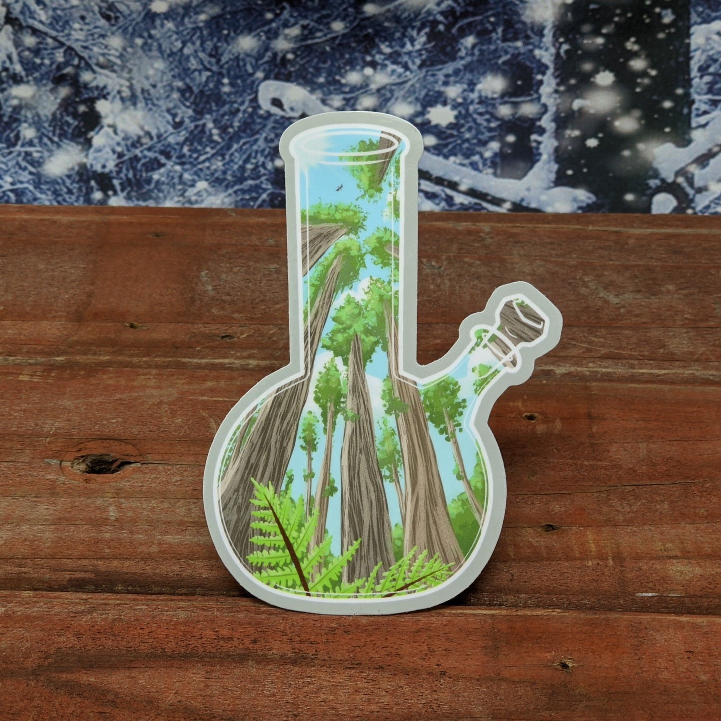 A redwood background with a snowy backdrop. On the wood is a vinyl sticker of a bong with a forest floor perspective scene of redwood trees and ferns.