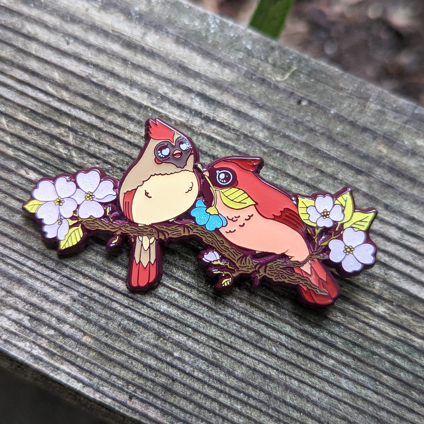 A photograph of a person holding an enamel pin in front of an orange flower. The enamel pin shows two cardinals on a dogwood tree surrounded by pink flowers. One of the cardinals is handing the other a special blue flower.