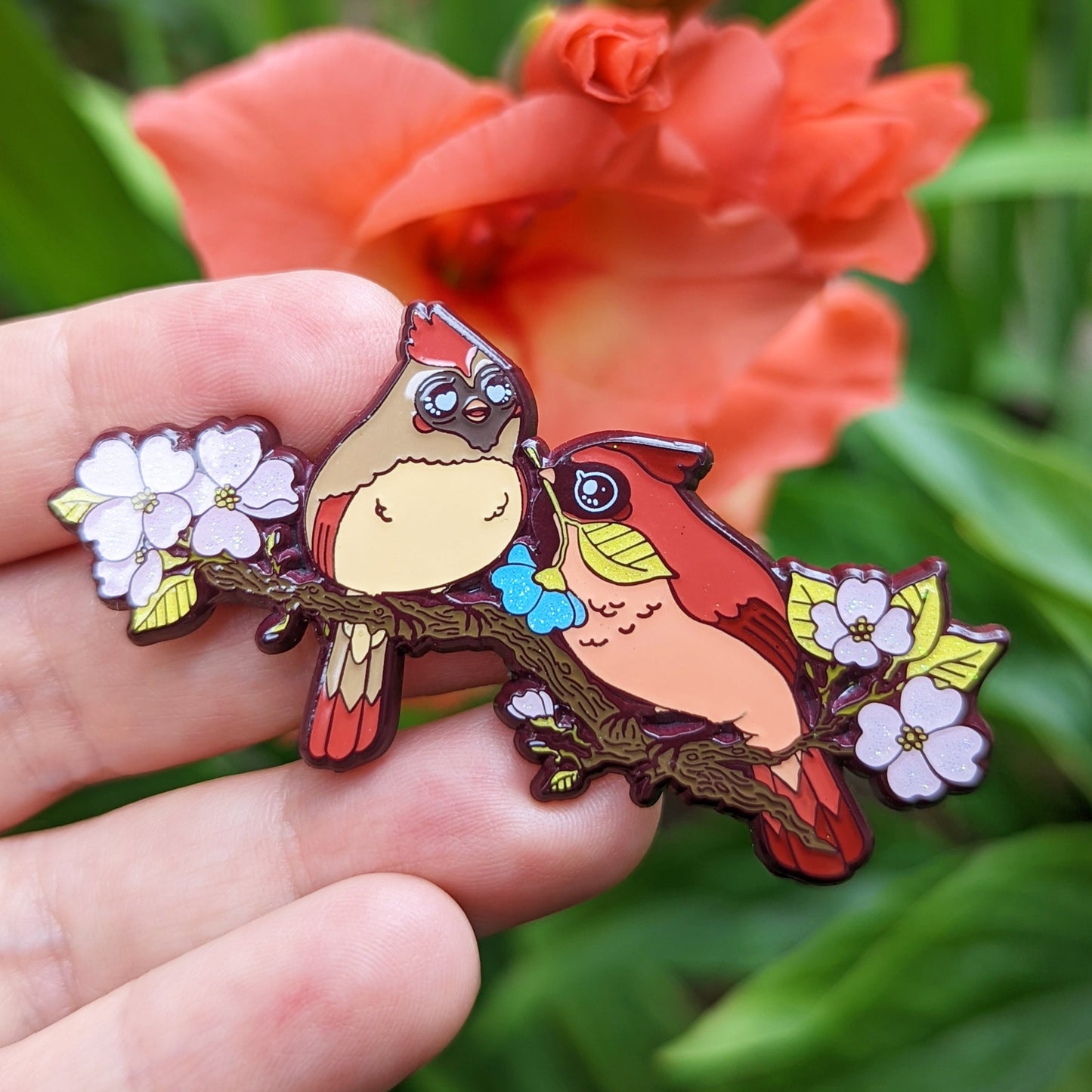 A photograph of a person holding an enamel pin in front of an orange flower. The enamel pin shows two cardinals on a dogwood tree surrounded by pink flowers. One of the cardinals is handing the other a special blue flower.