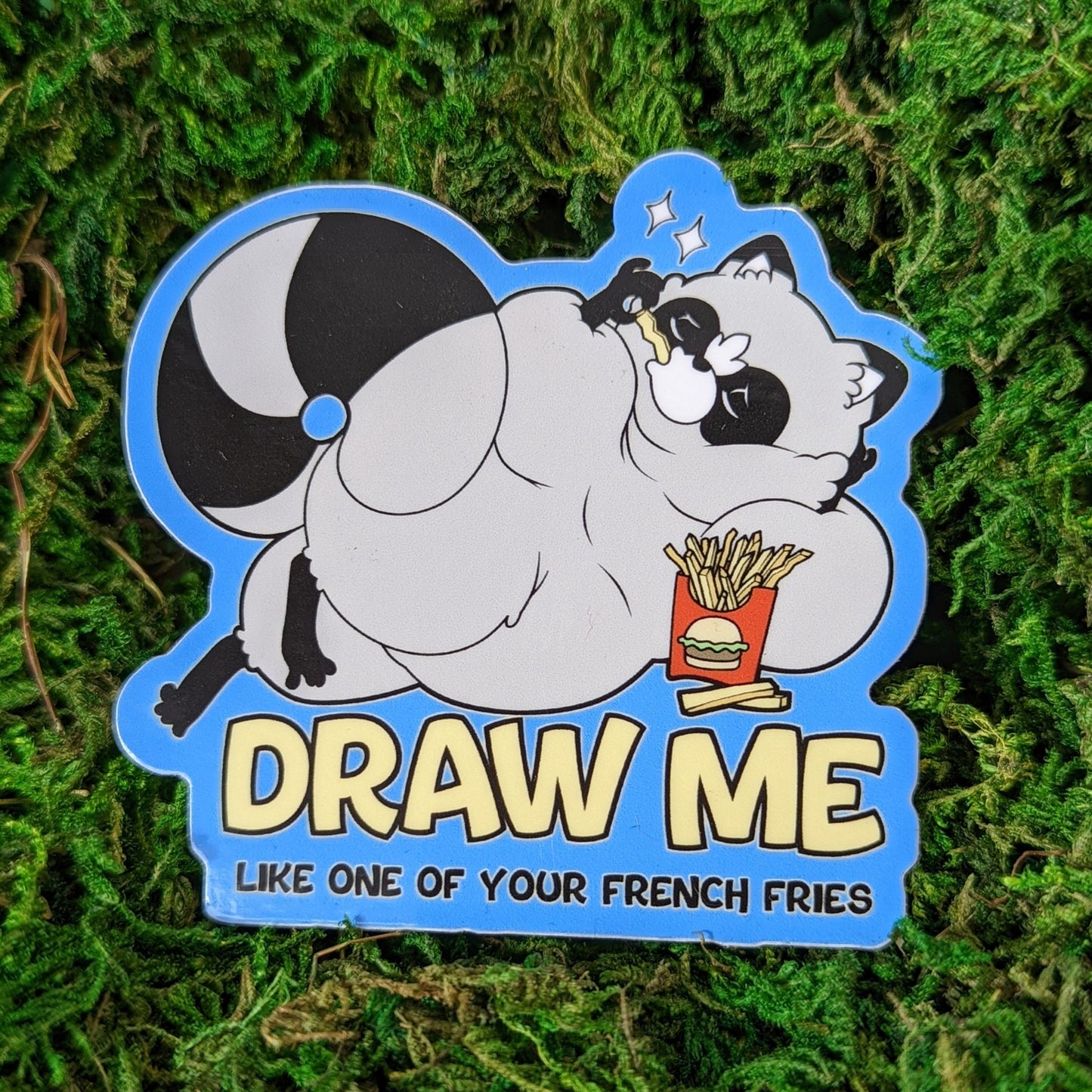 Photograph of a mossy background with a vinyl sticker of a raccoon with laid out while eating french fries. Under the raccoon it says, “Draw me like one of your french fries.”