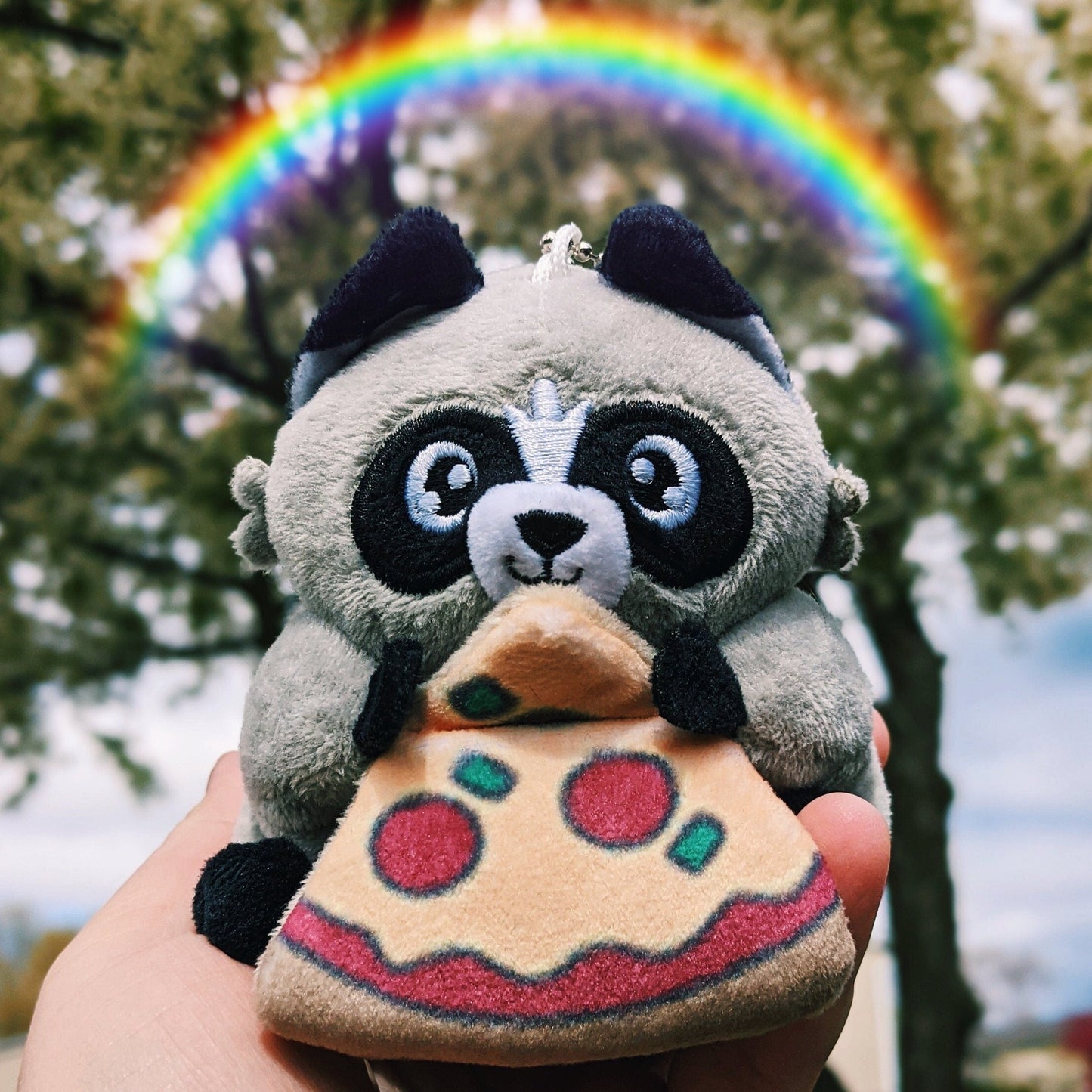 A photograph of Chaps the Trash Panda Plush. Over chaps is a rainbow. The Chaps plush is a small handsize raccoon plush toy holding a slice of pizza.