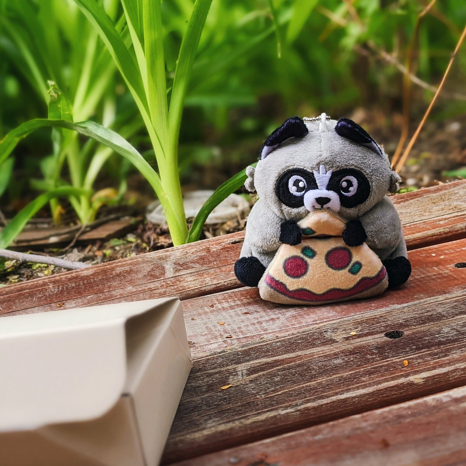 A photograph of Chaps the Trash Panda Plush. Chaps is sitting on a wood deck looking at a pizza box. The Chaps plush is a small handsize raccoon plush toy holding a slice of pizza.