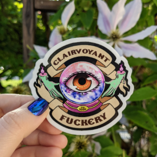 A photograph of a vinyl sticker being held in front of a clematis plant. The sticker is of a crystal ball with an eye in the middle, two candle hands are floating around it. There’s a scroll banner around the crystal ball that says “Clairvoyant to your fuckery.”